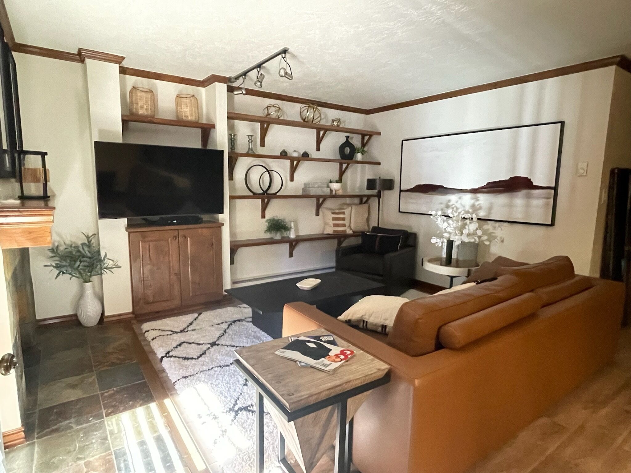A living room with a brown leather sofa, a TV on a wooden cabinet, a coffee table, and a bookshelf with decorative items.