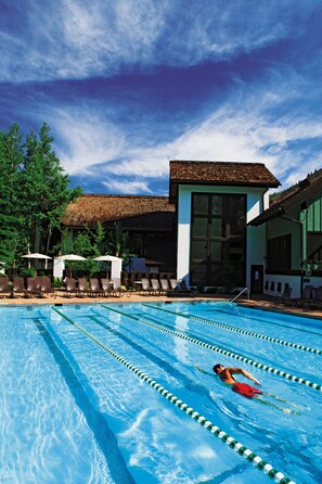 A swimming pool with lane dividers, a person swimming, lounge chairs, and a building with large windows and a thatched roof in the background. - Vail Racquet Club Mountain Resort (Vail)