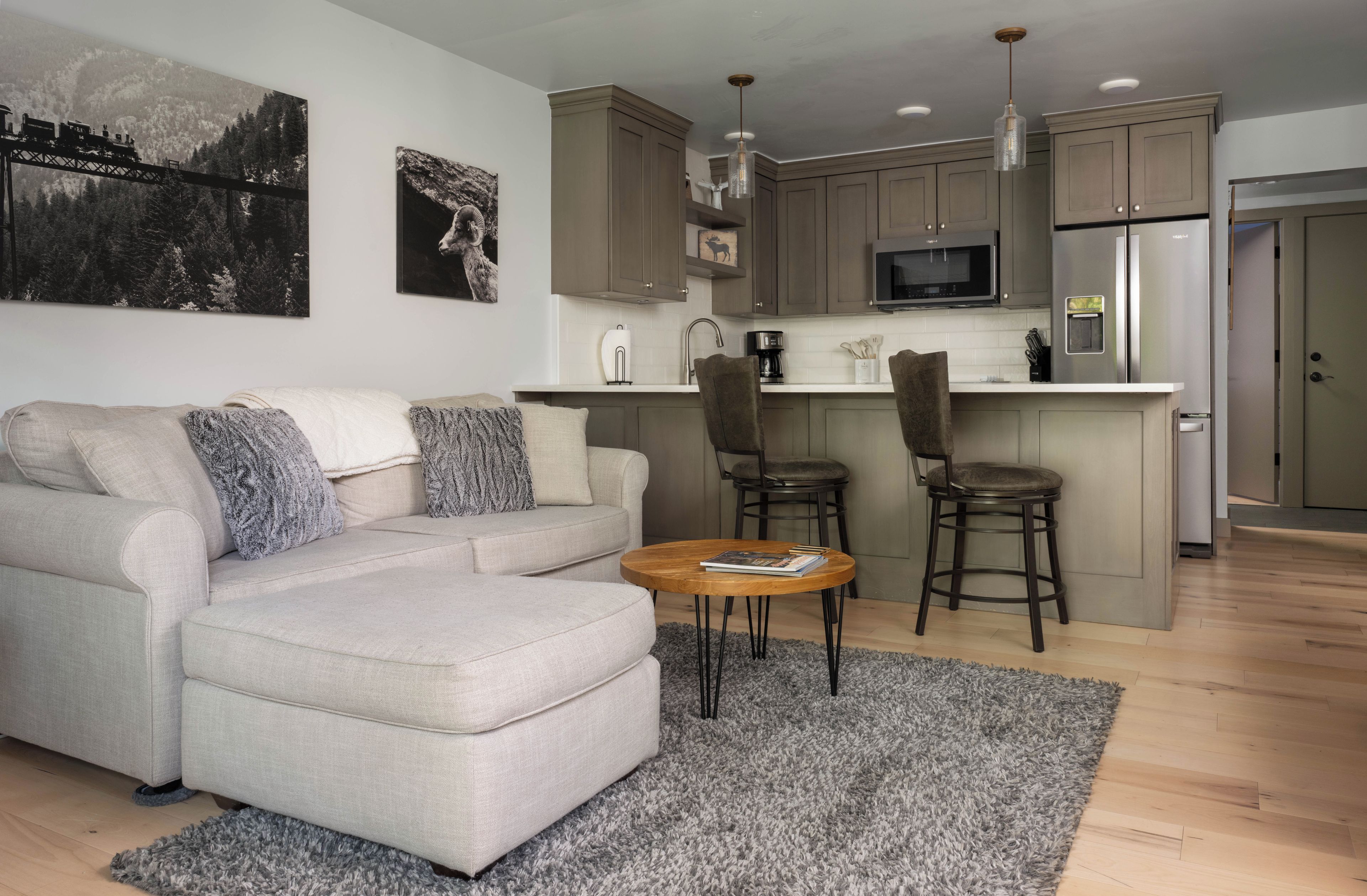 A modern living room with a sofa, a coffee table, and a kitchen area with a microwave, refrigerator, and bar stools.