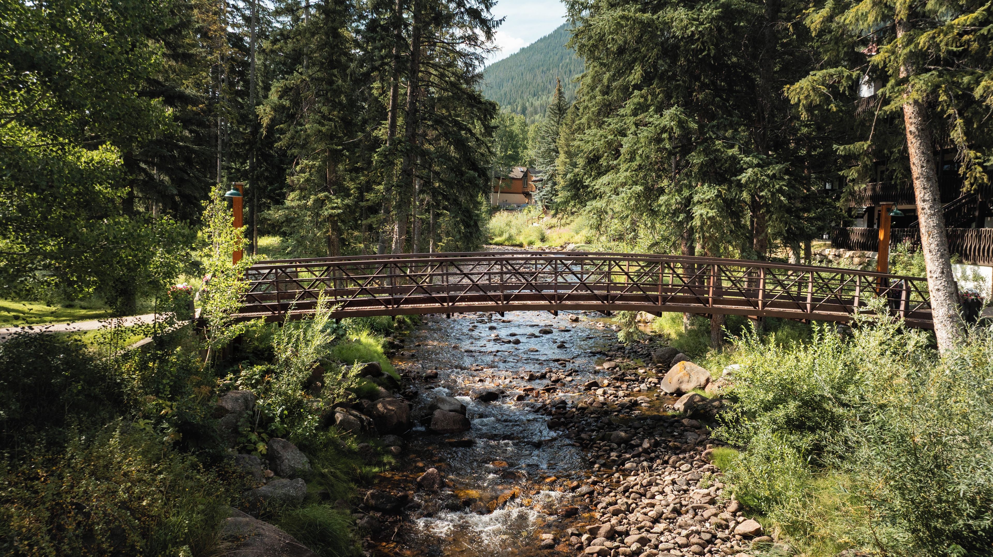 A wooden bridge over a rocky stream in a forested area with a building in the background.