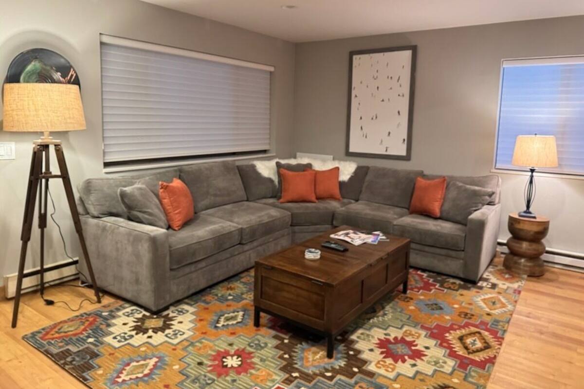 Un salon avec un canapé d’angle gris, une table basse en bois et un tapis coloré.