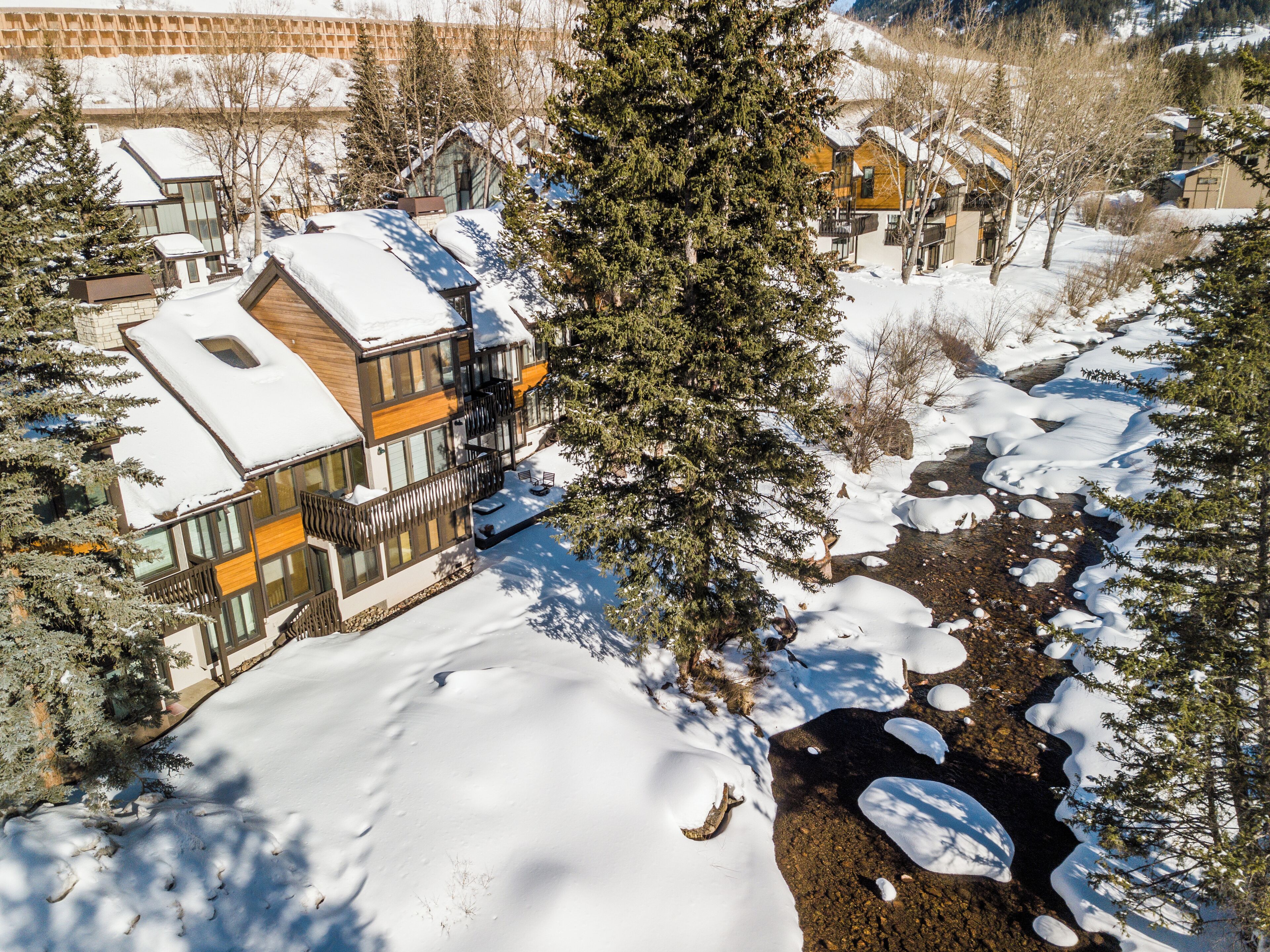 Ein modernes, zweistöckiges Haus mit schneebedecktem Dach und einem Balkon, umgeben von Bäumen und einem kleinen Bach.