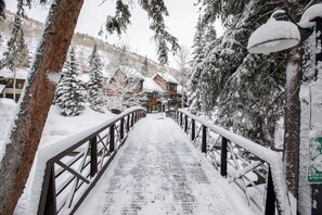 Uma ponte de madeira coberta por neve que leva a um edifício com telhado inclinado, cercado por árvores carregadas de neve.