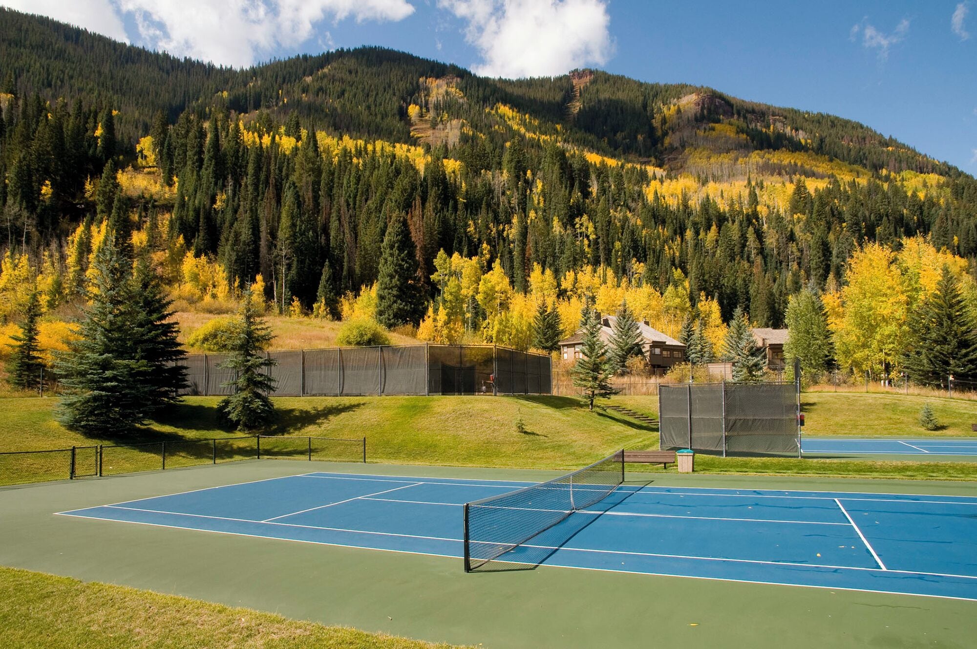 Un court de tennis en plein air, doté d’un filet et entouré d’une clôture, avec une montagne boisée en arrière-plan.