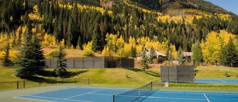An outdoor tennis court with a net, surrounded by a fence, and a forested mountain in the background.