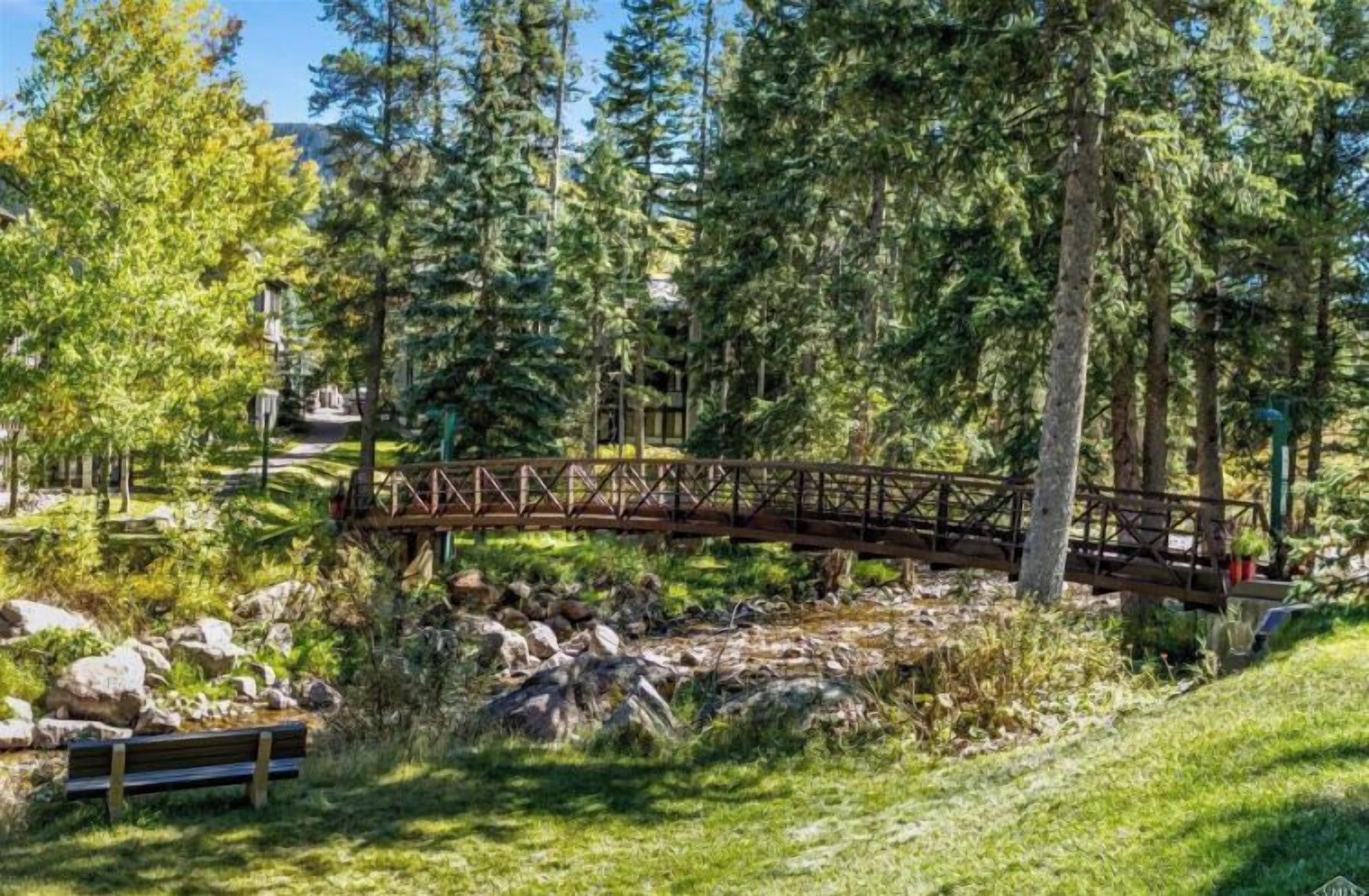 A wooden bridge over a rocky stream in a forested area with a bench and trees.