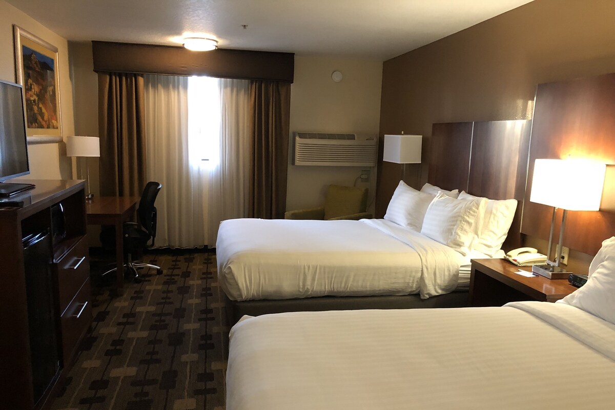 A hotel room with two beds, a desk, a chair, a lamp, and a painting on the wall.