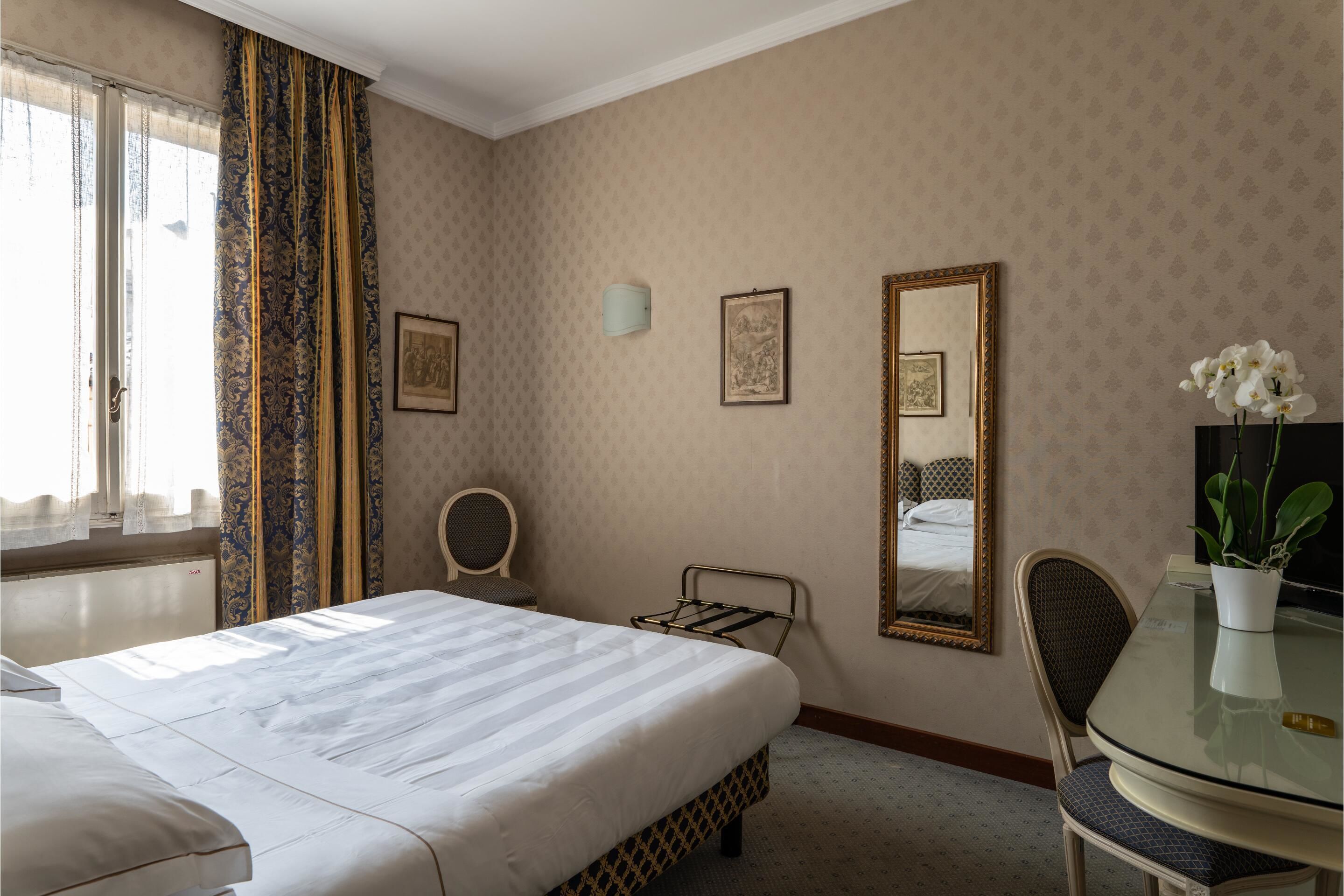 A hotel room with a large bed, a small table, a chair, a mirror, and a potted plant.