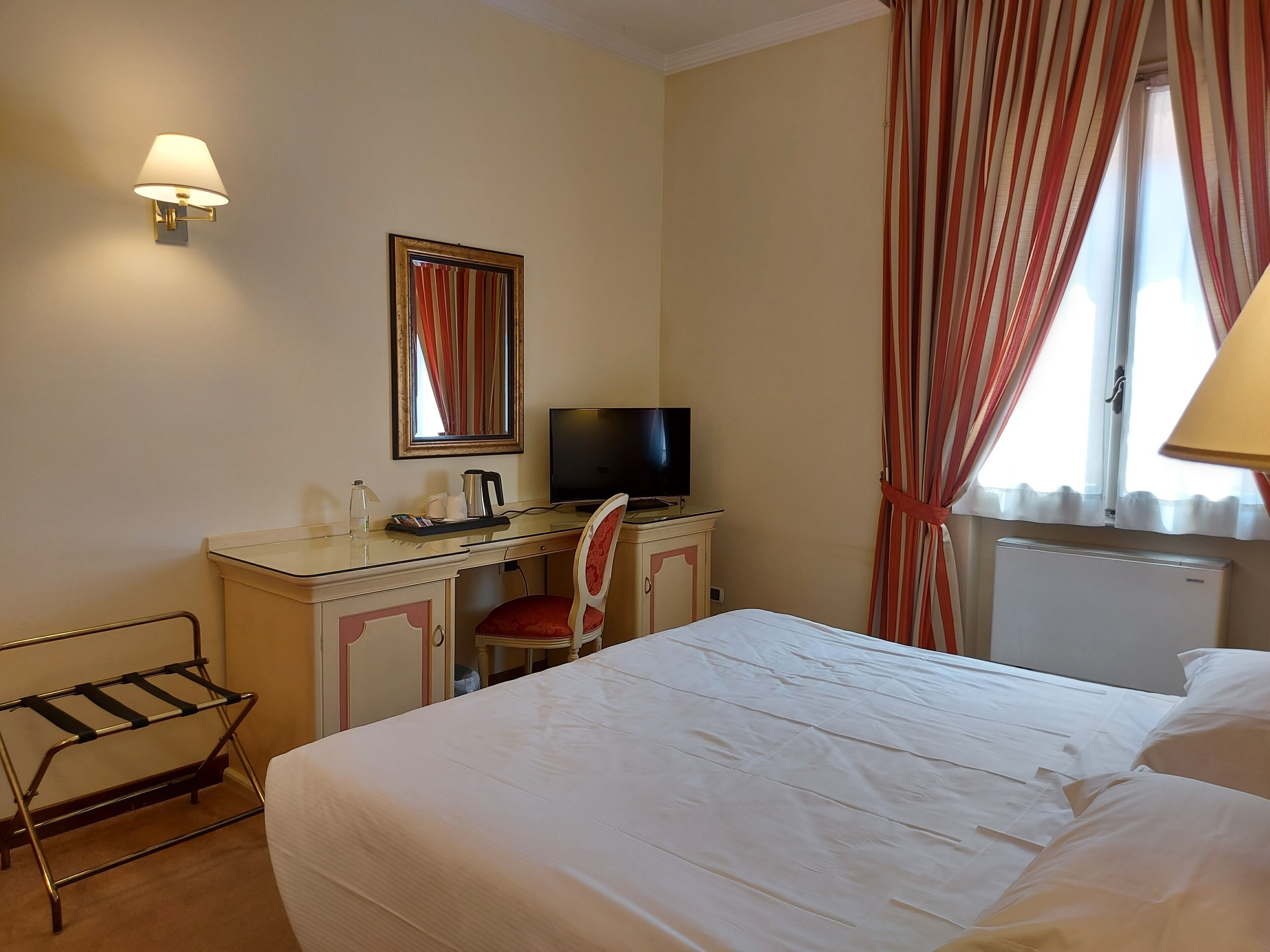 A hotel room with a bed, a desk with a computer, a chair, a TV, and a radiator.