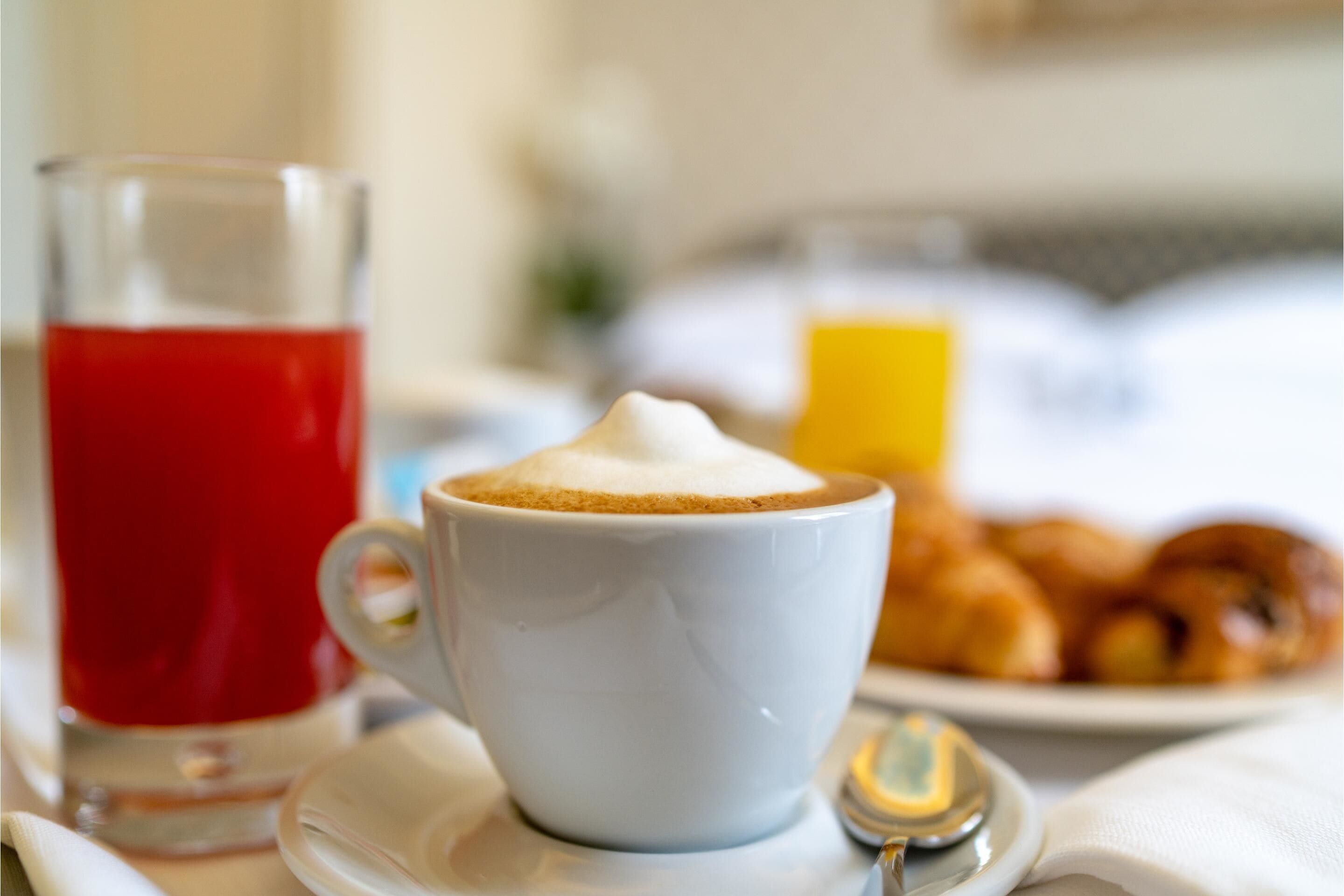 En vit kaffekopp med skum på toppen, ett glas med en röd dryck och en tallrik med croissanter.