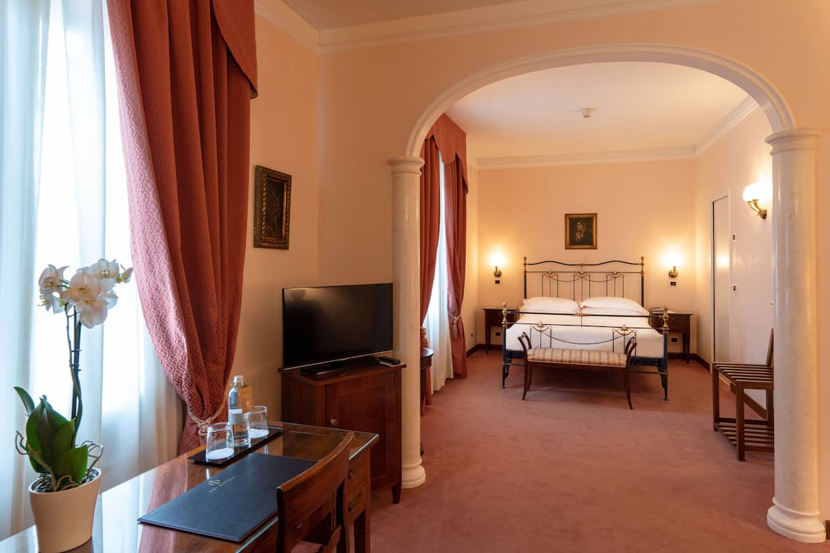 Habitación de hotel con cama, escritorio con televisor, una silla, planta en maceta y vista a través de un arco.