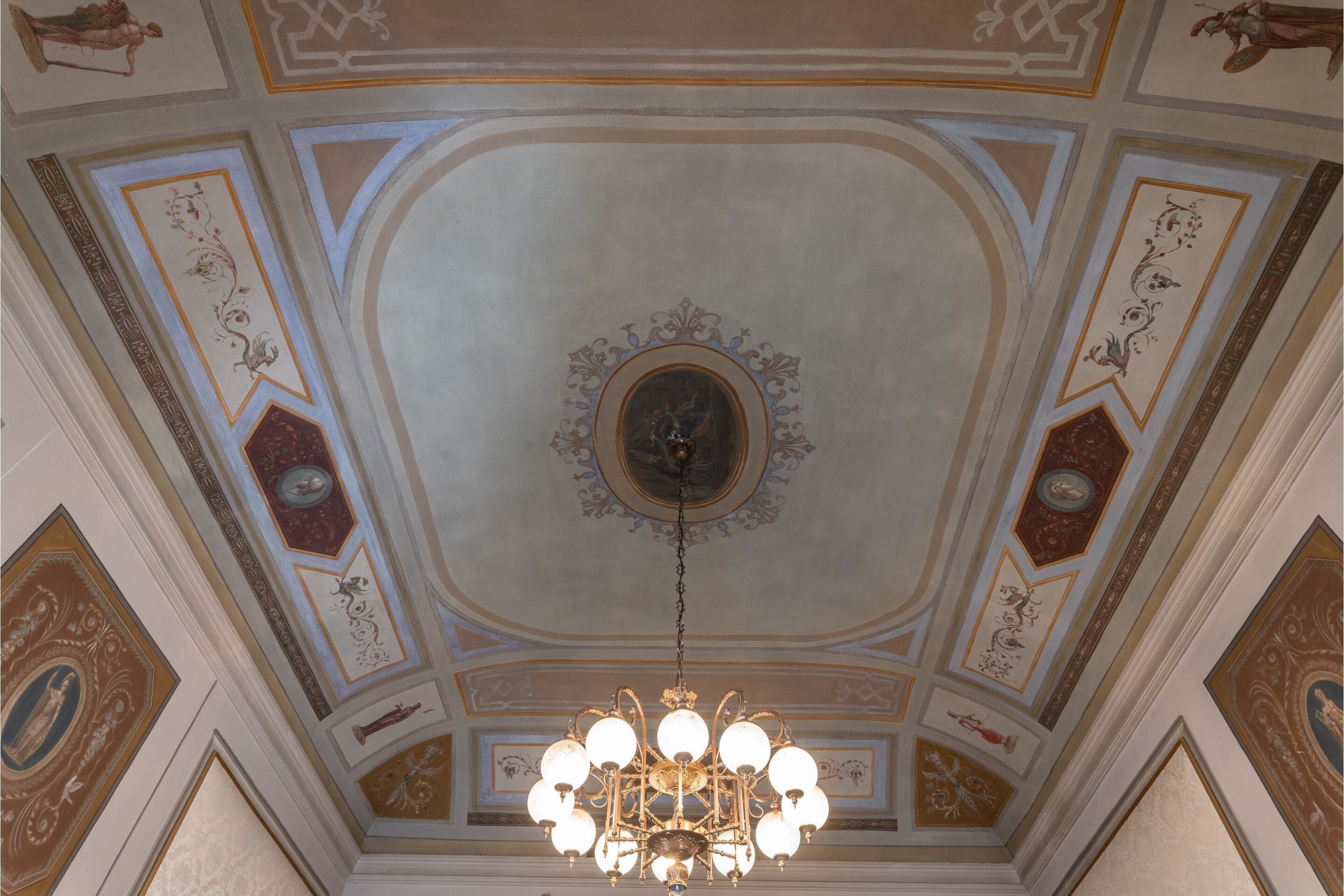 Un soffitto con decorazioni elaborate, un lampadario centrale e opere d'arte incorniciate.
