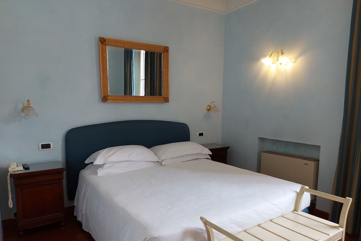 Una cama bien tendida con cabecera, una mesita de noche con lámpara y un espejo fijado a la pared.