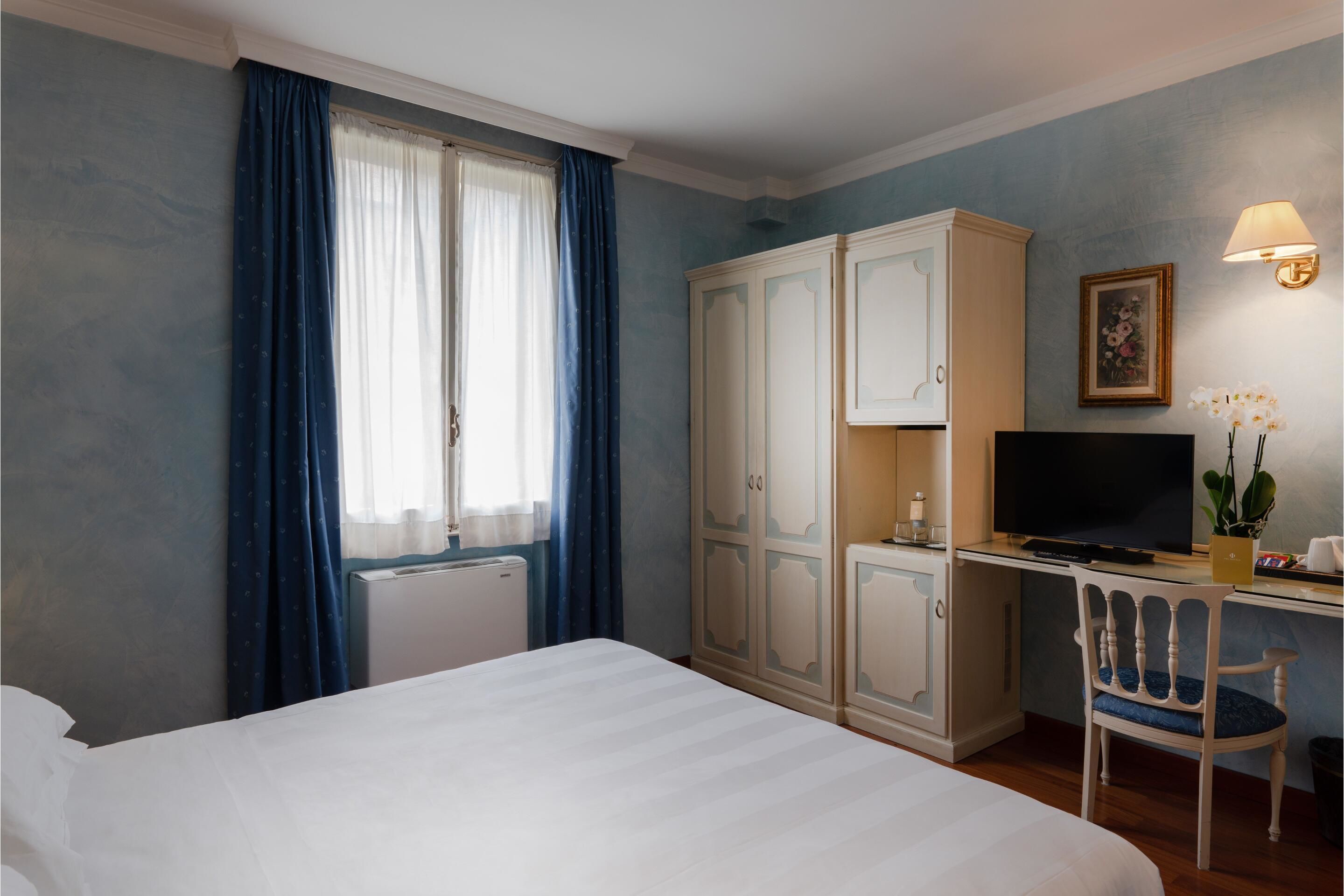 A hotel room with a bed, a desk with a TV, a wardrobe, and a window with curtains.