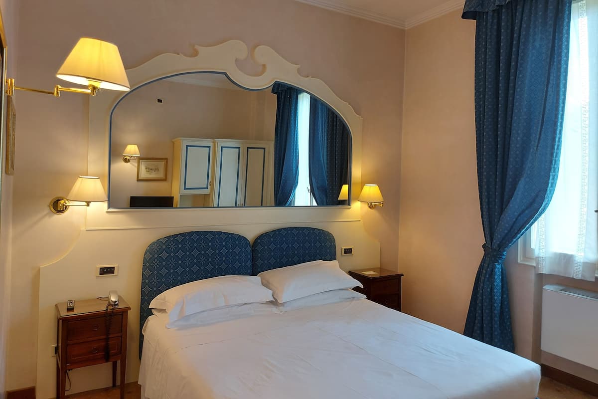 Habitación de hotel con una cama grande, dos mesitas de noche, un espejo y una ventana con cortinas.
