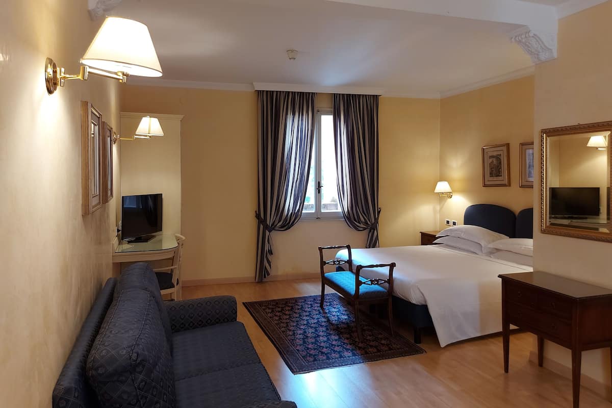 Una habitación de hotel con cama, sofá, escritorio y televisión, además de una ventana con cortinas.