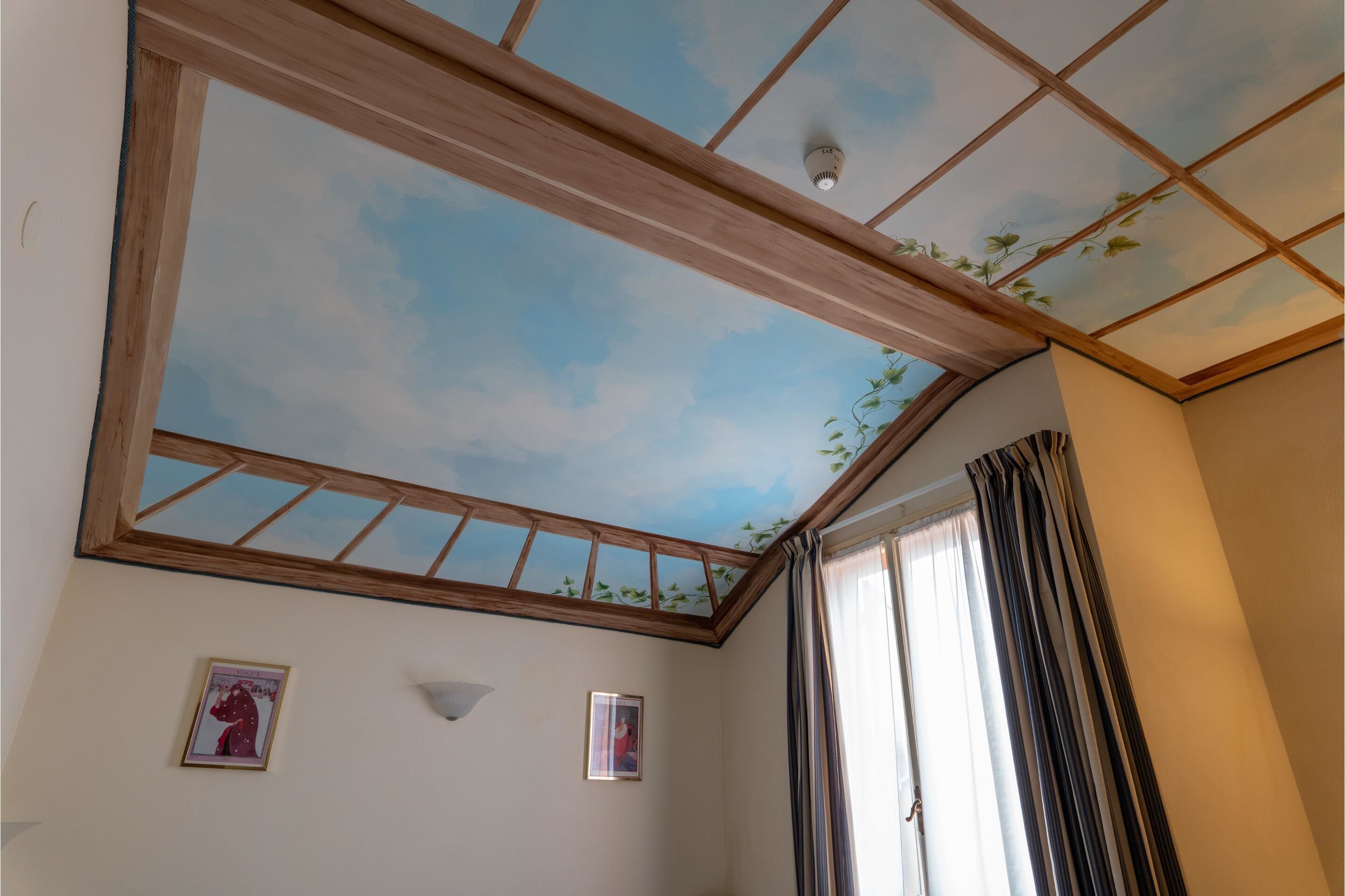 Una stanza con lucernario, travi in legno, soffitto affrescato con cielo dipinto e due quadri appesi al muro.