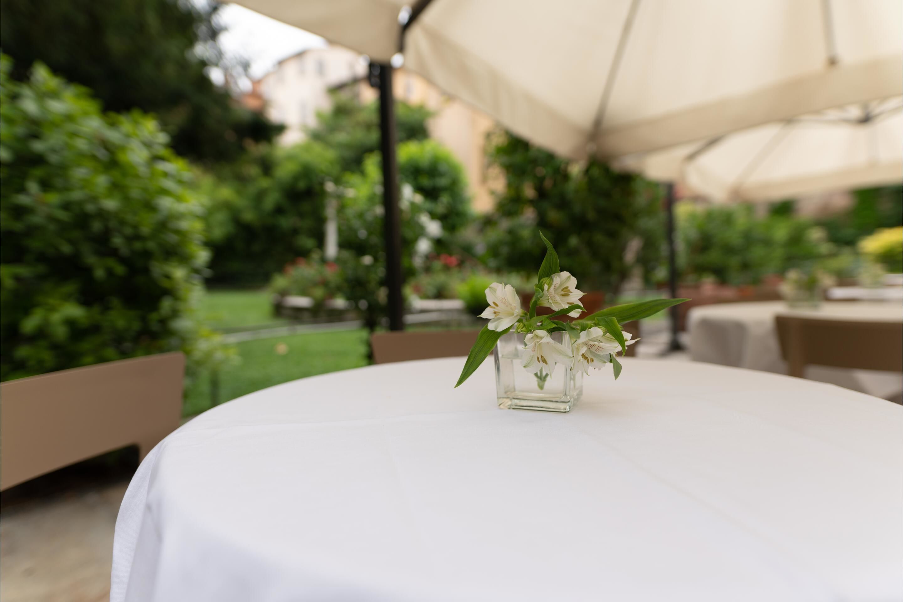 A round table with a white tablecloth, a glass vase with white flowers, and a beige umbrella in the background.