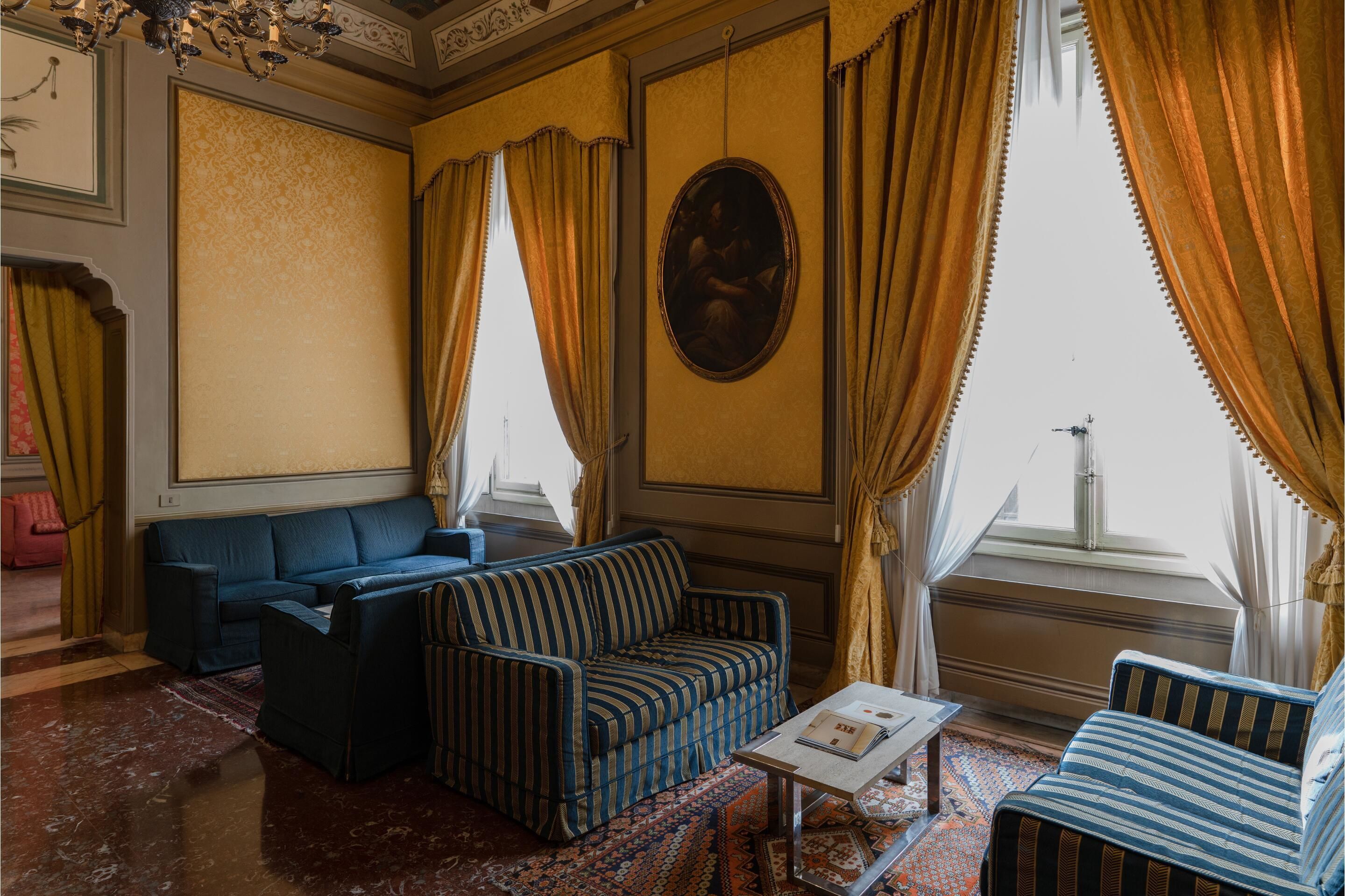 A room with a blue sofa, a striped armchair, and a small table. There is a large window with curtains, and a painting on the wall.
