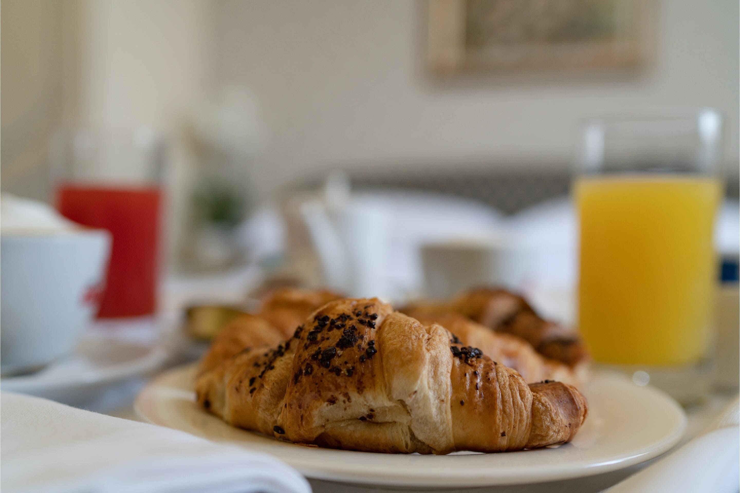 Une table mise pour le petit-déjeuner avec des croissants sur une assiette, un verre de jus d’orange et une tasse rouge.