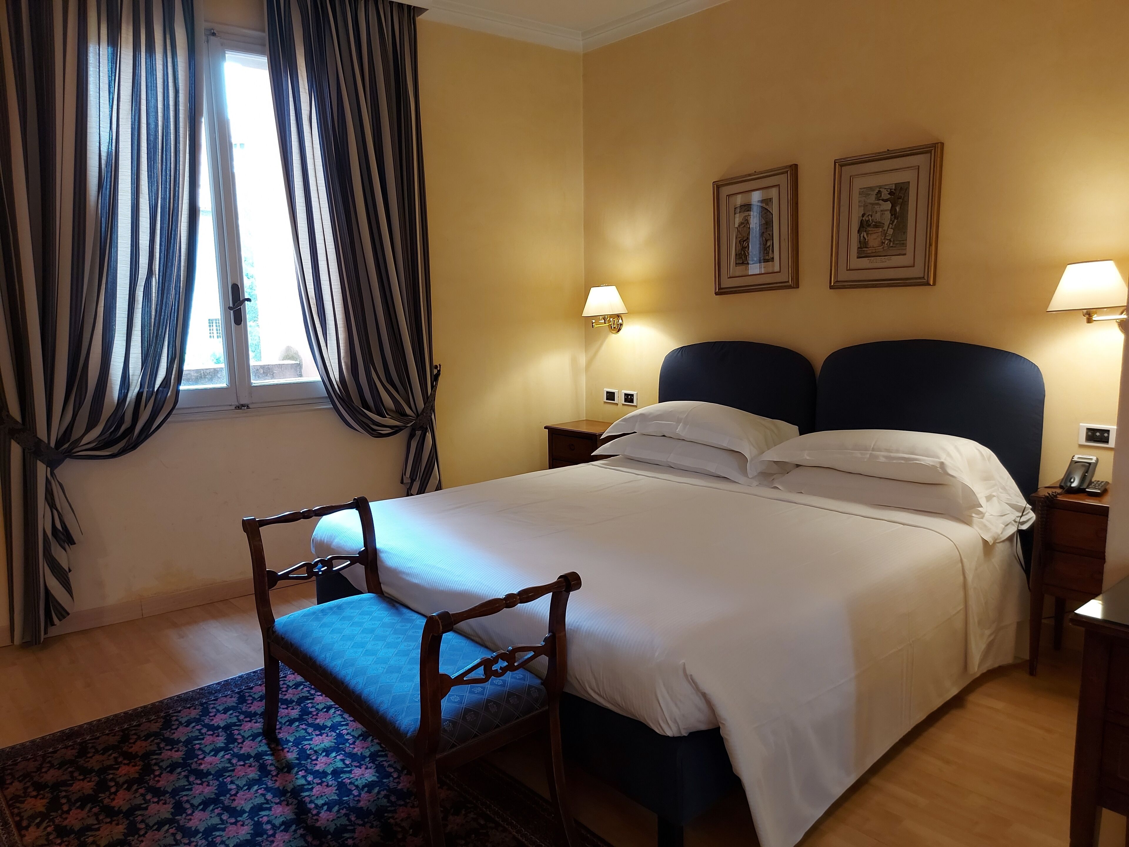 A hotel room with a bed, two bedside lamps, a nightstand, a chair, a window with curtains, and two framed pictures on the wall.