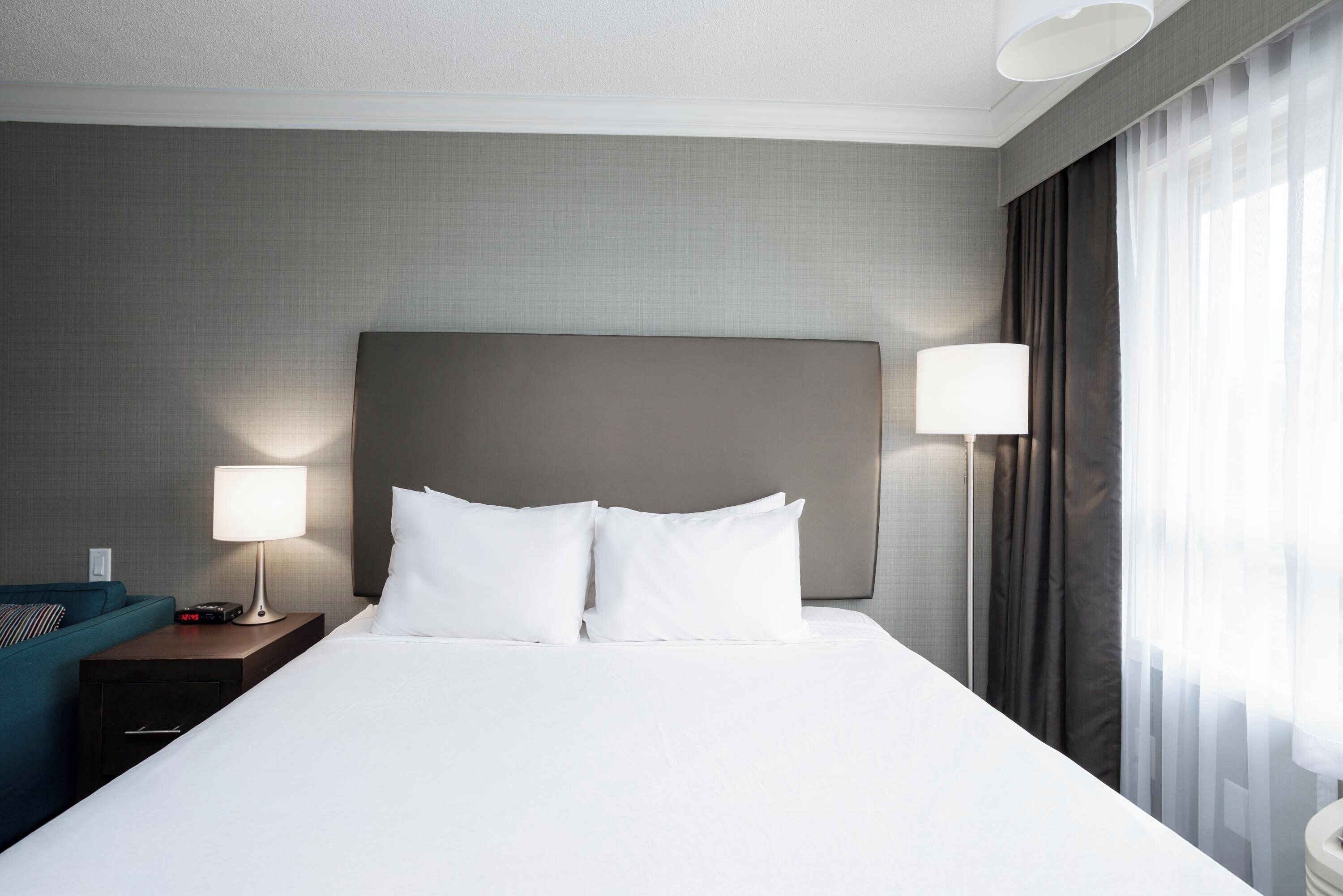 A hotel room with a large bed, a nightstand with a lamp, a blue chair, and a window with curtains.