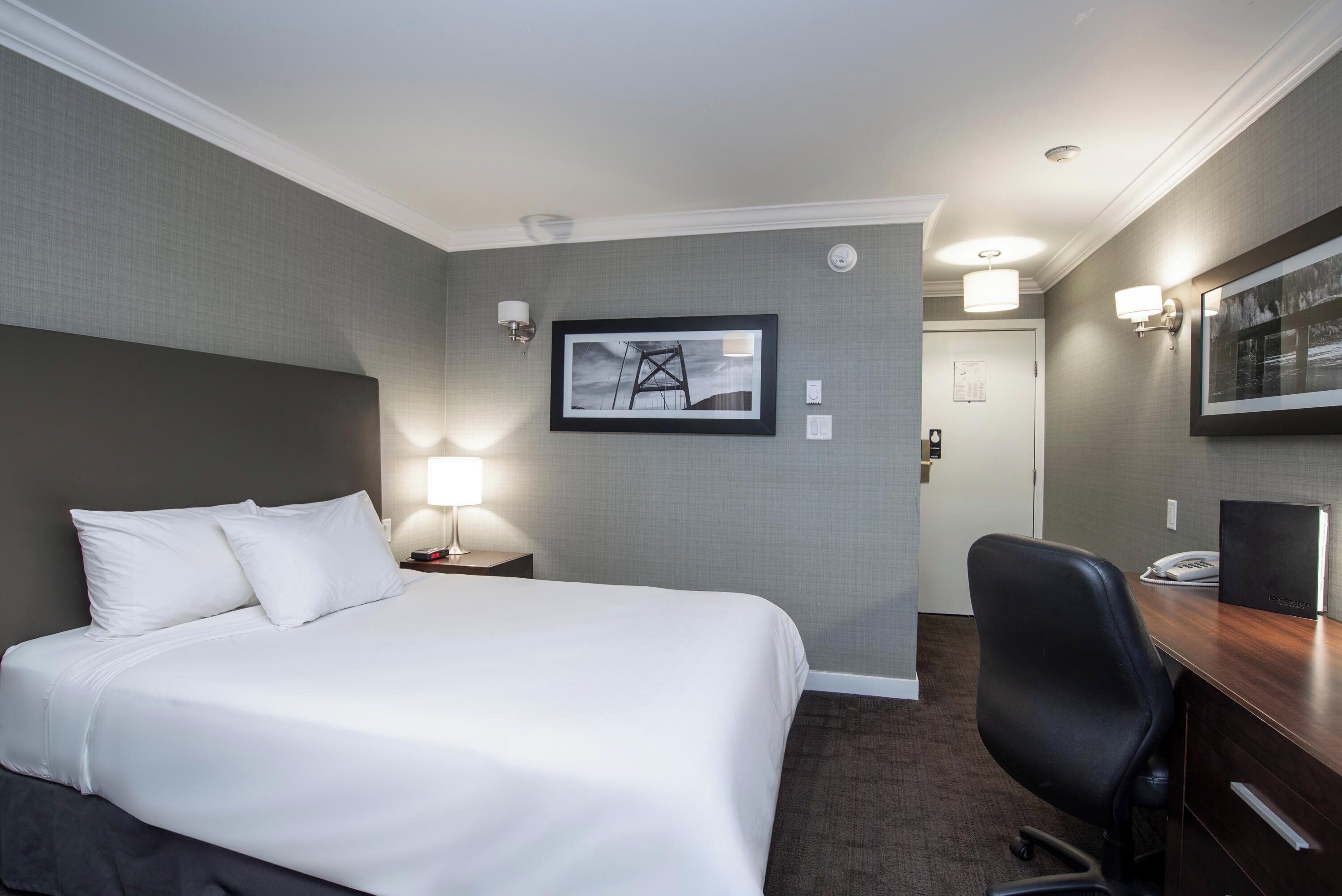 A hotel room with a large bed, a desk, and a chair. There is a framed picture on the wall and a lamp on the nightstand.