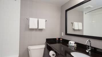 Un baño moderno con inodoro, lavamanos y espejo. Hay toallas colgadas en un perchero y un rollo de papel higiénico en la encimera.