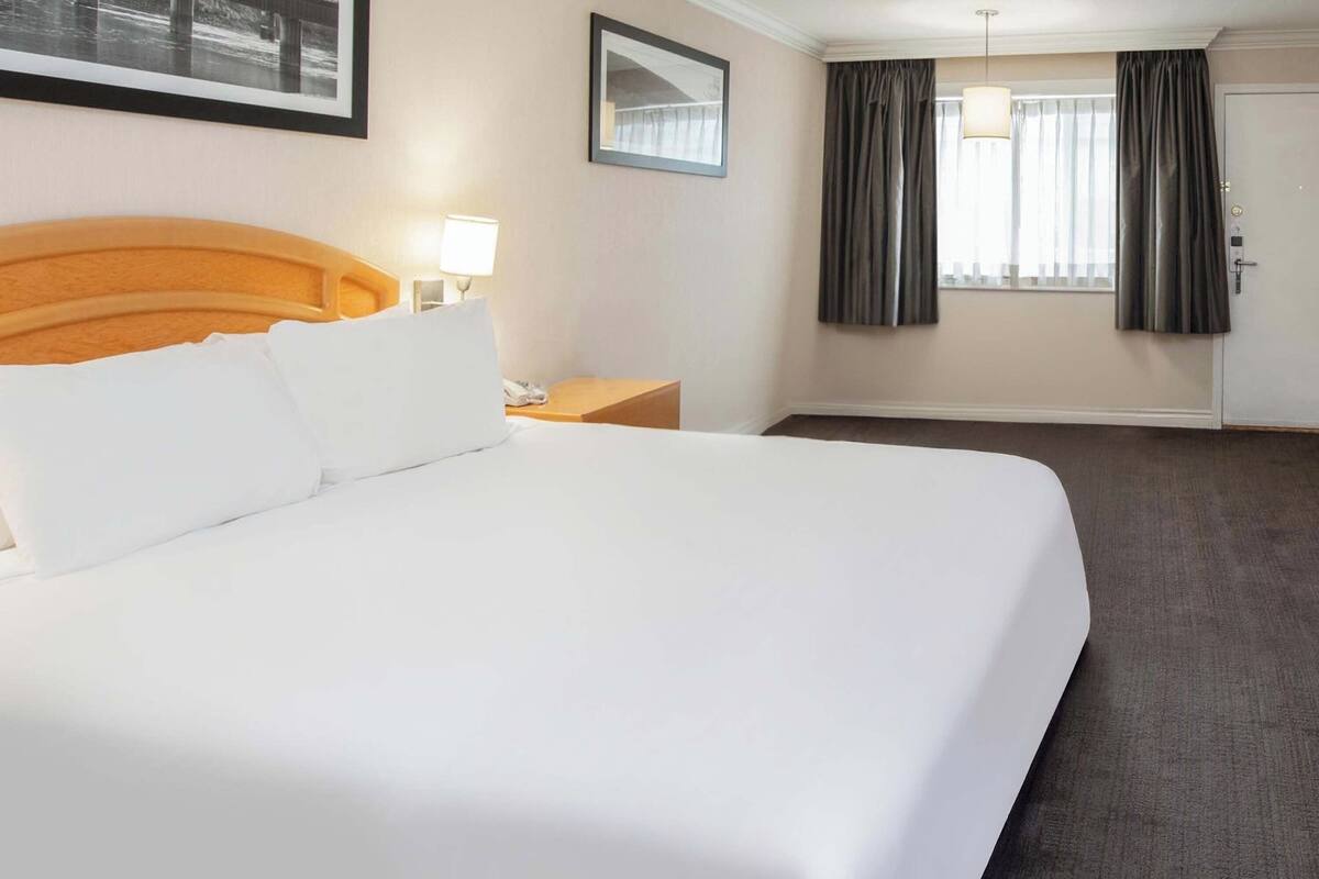 A hotel room with a large bed, a nightstand, a lamp, a framed picture on the wall, and a window with curtains.