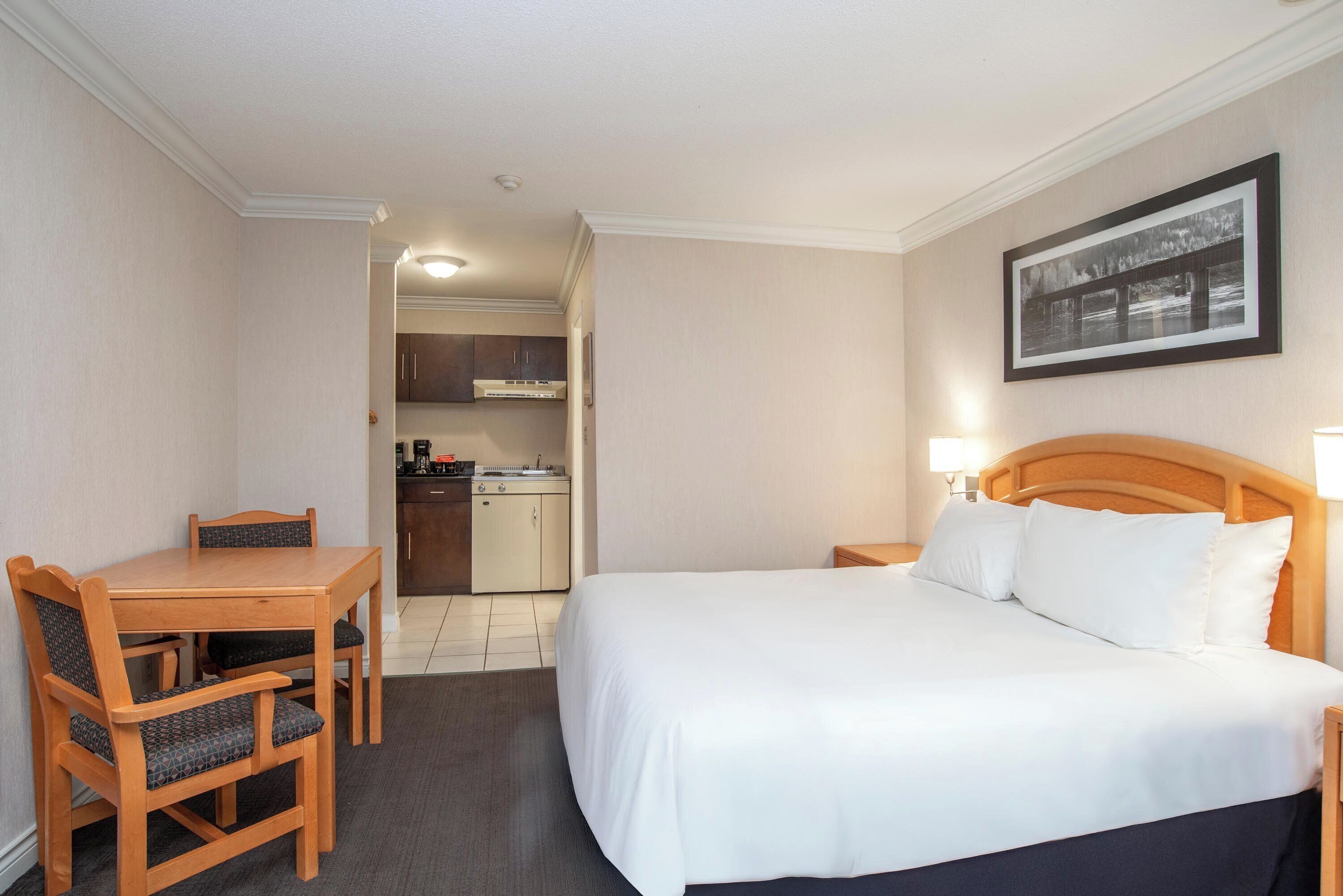 A hotel room with a bed, a desk, and a chair. There is a kitchenette in the background.