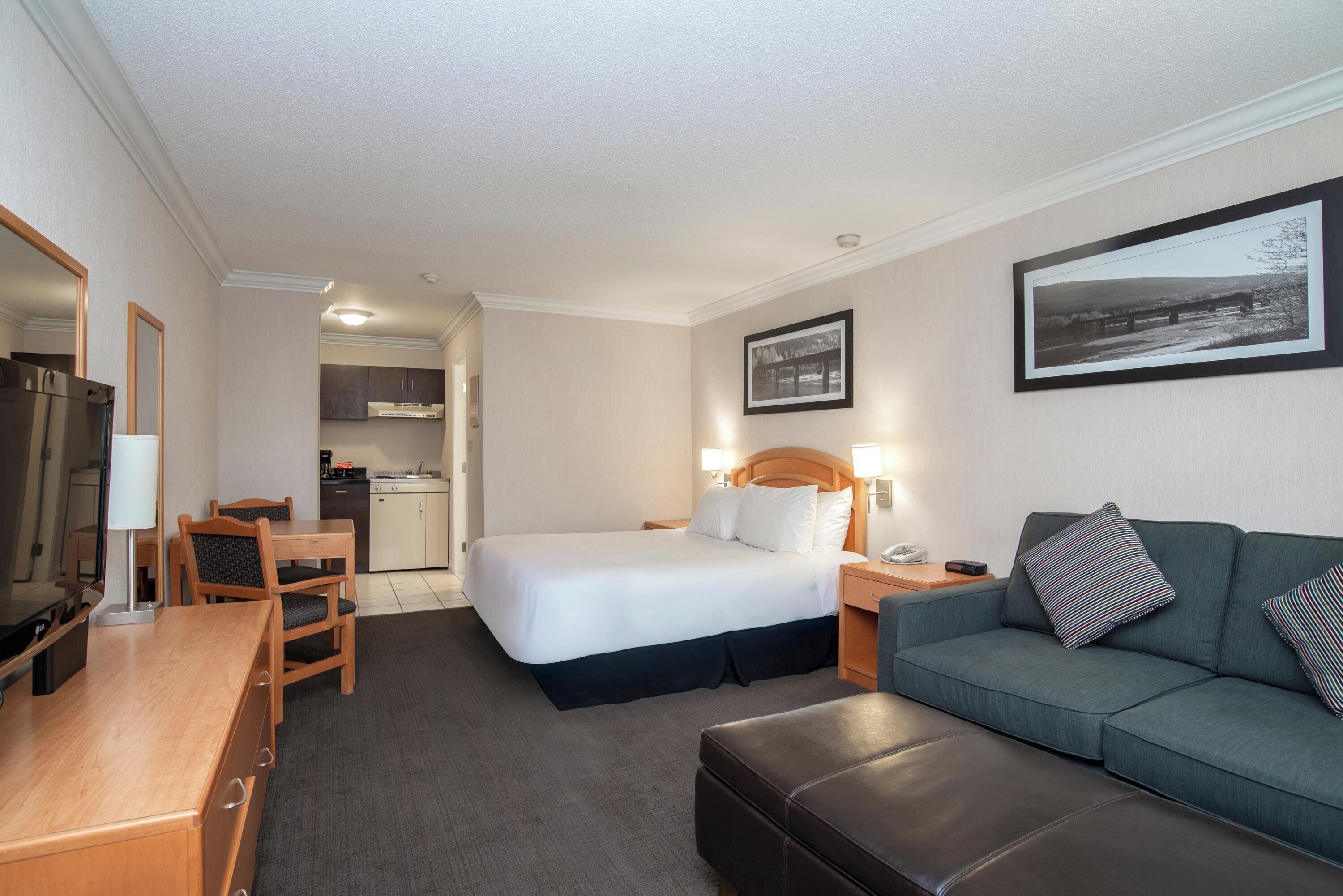 A hotel room with a bed, a sofa, a desk, a chair, a television, a kitchen area, and a framed picture on the wall.