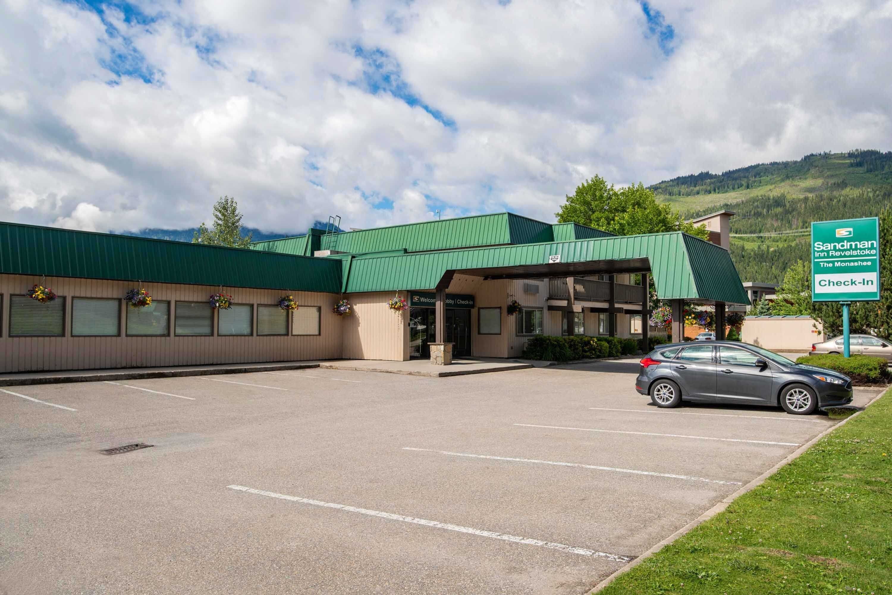 A Sandman Inn Revelstoke building with a green roof, a parking lot, and a car parked in front.