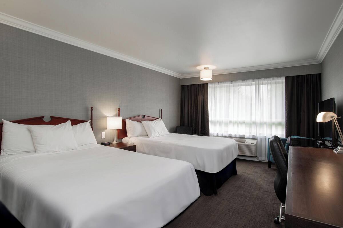 A hotel room with two beds, a desk, a chair, a lamp, a nightstand, a window with curtains, and an air conditioning unit.