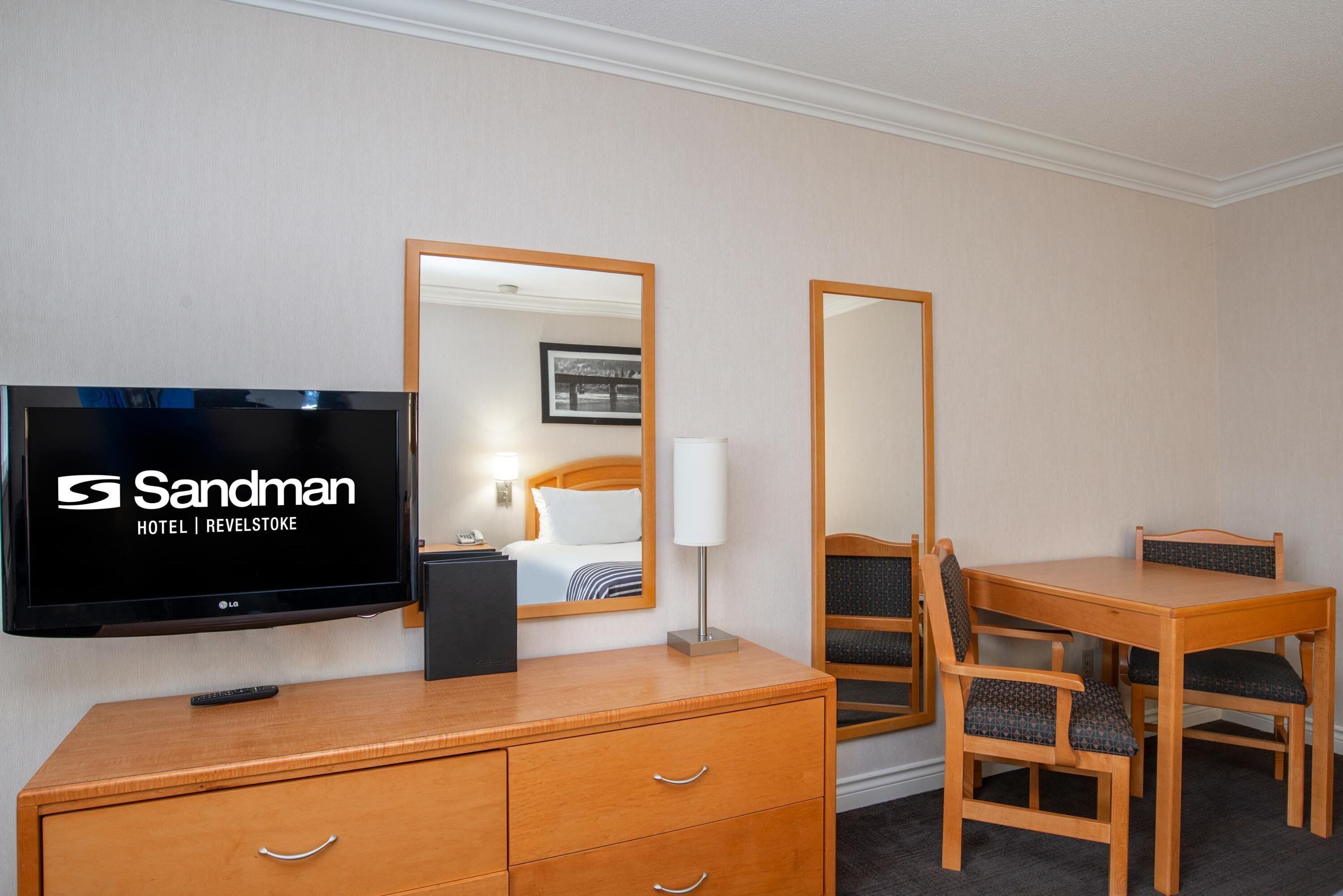 A hotel room with a flat-screen TV, wooden furniture, a mirror, and a bed with a white headboard.