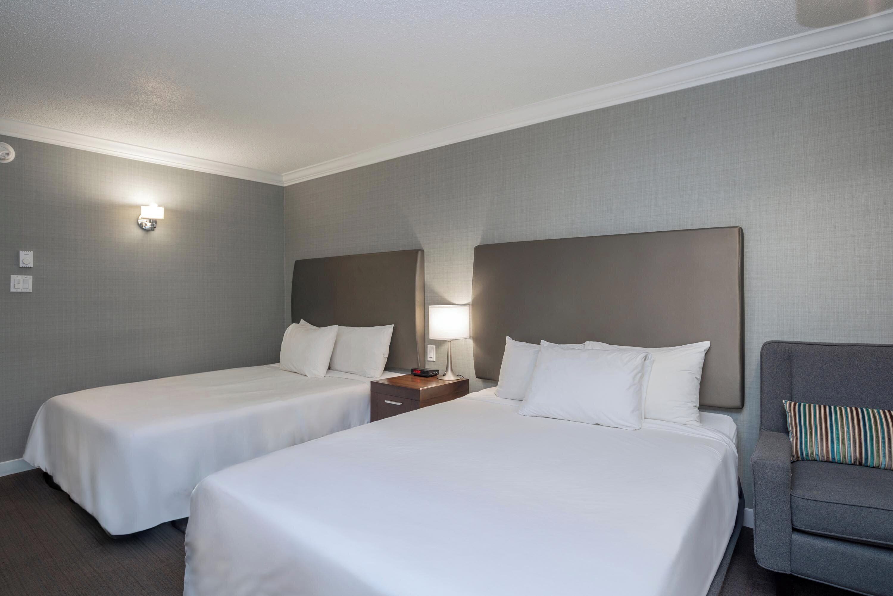 A hotel room with two single beds, a nightstand with a lamp, a gray armchair, and a wall-mounted light fixture.