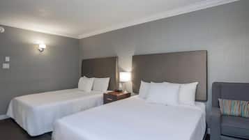 Una habitación de hotel con dos camas individuales, una mesita de noche con lámpara, un sillón gris y un aplique de pared.