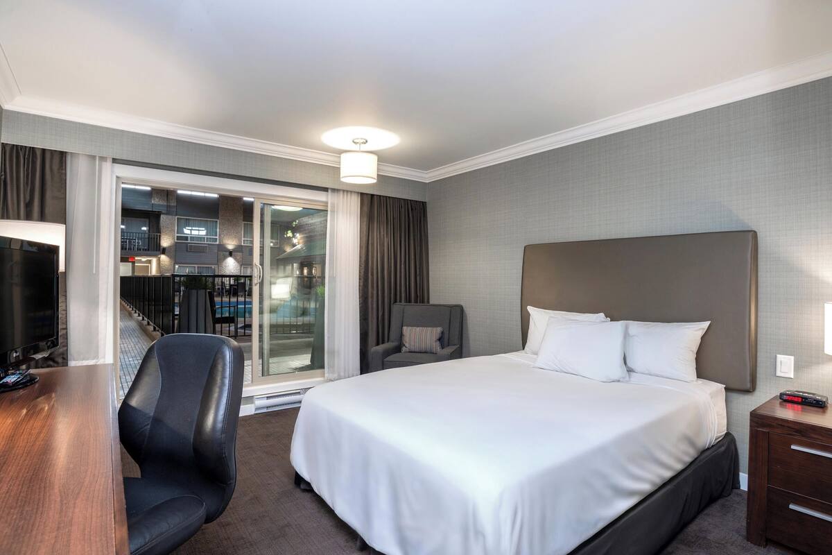 A hotel room with a large bed, a desk, a chair, a TV, and a view of the outside.