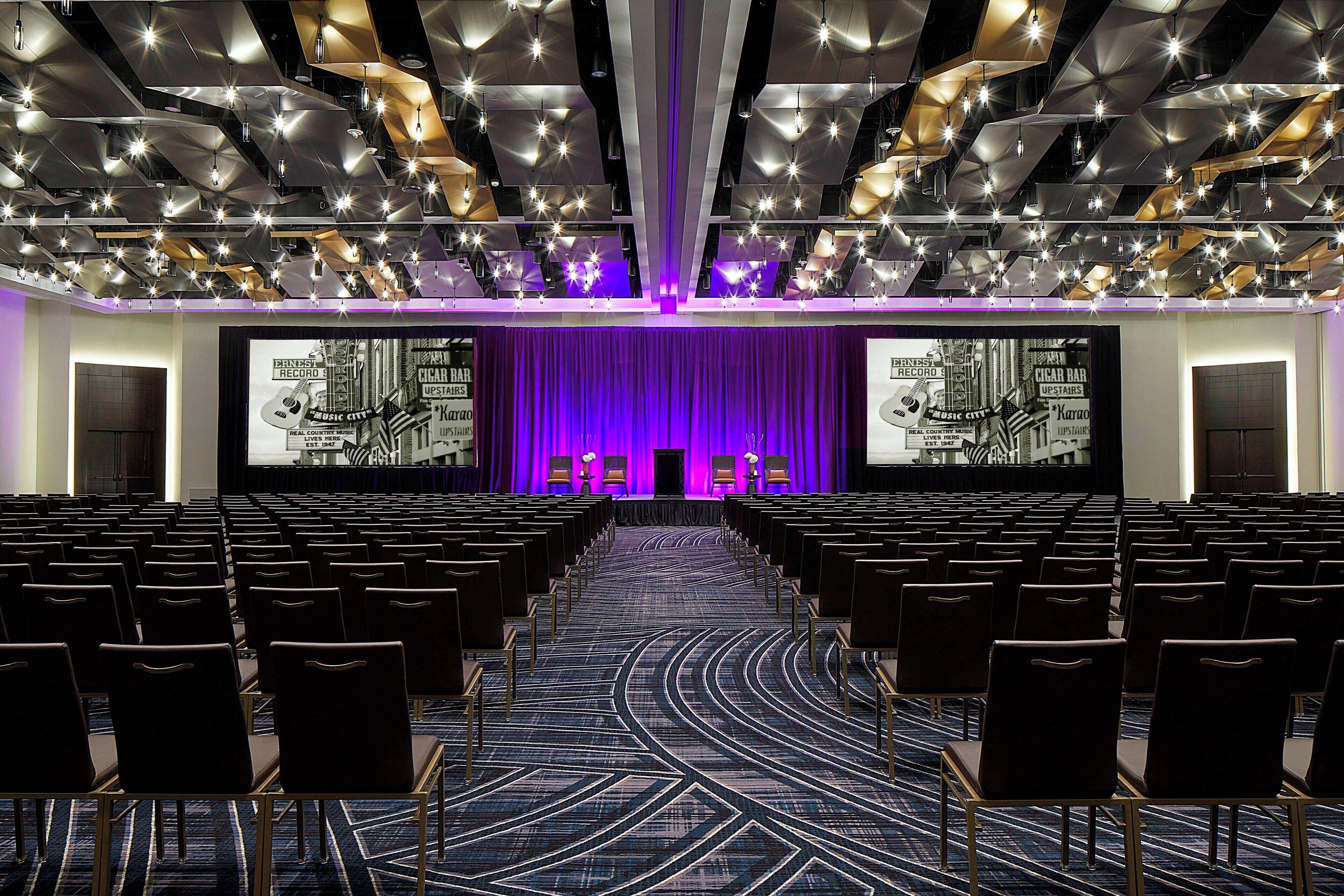 Un salón de conferencias con filas de sillas, un escenario con cortina púrpura y una pantalla grande que muestra un gráfico.