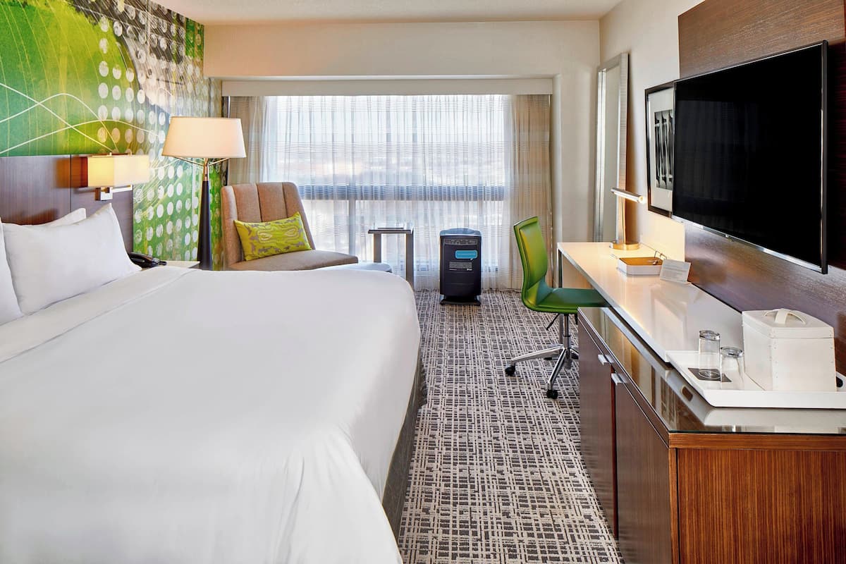 Habitación de hotel con una cama grande, un escritorio con televisor de pantalla plana, una silla y vistas a la ciudad desde la ventana.