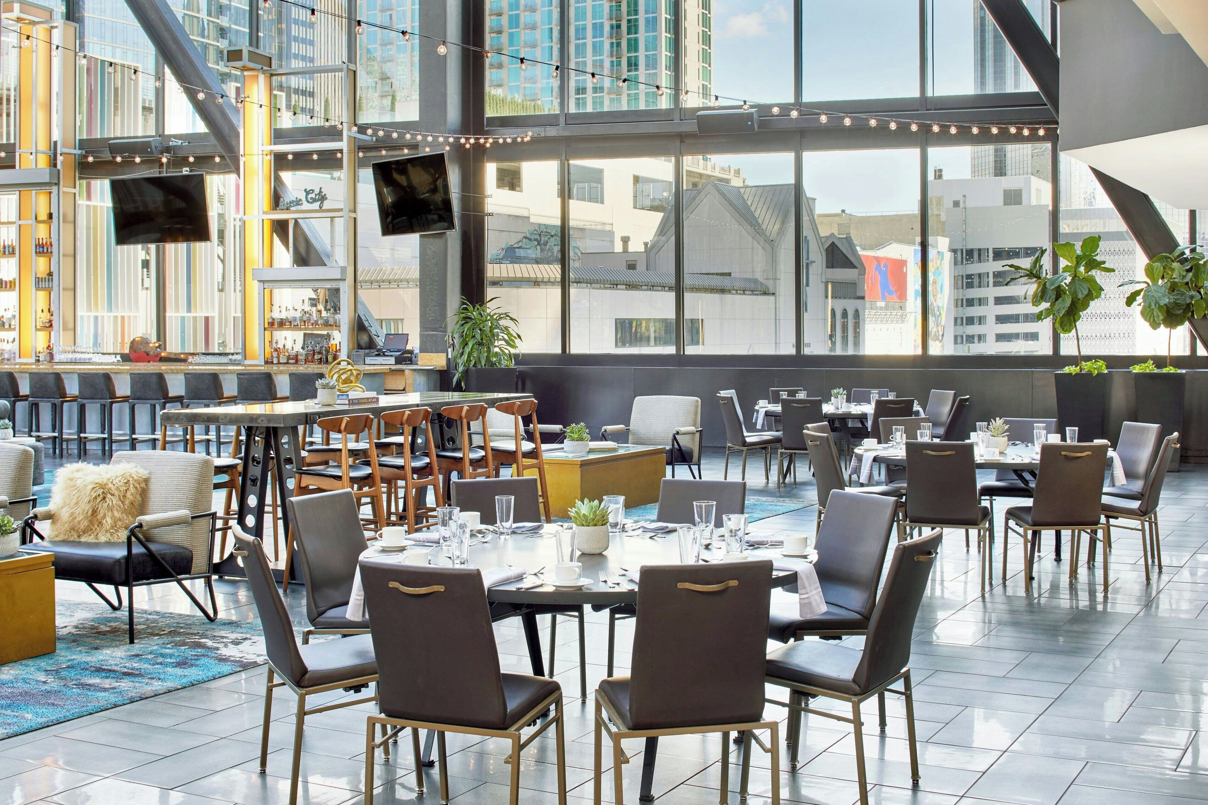 A modern dining area with multiple tables and chairs, large windows offering city views, and indoor plants.