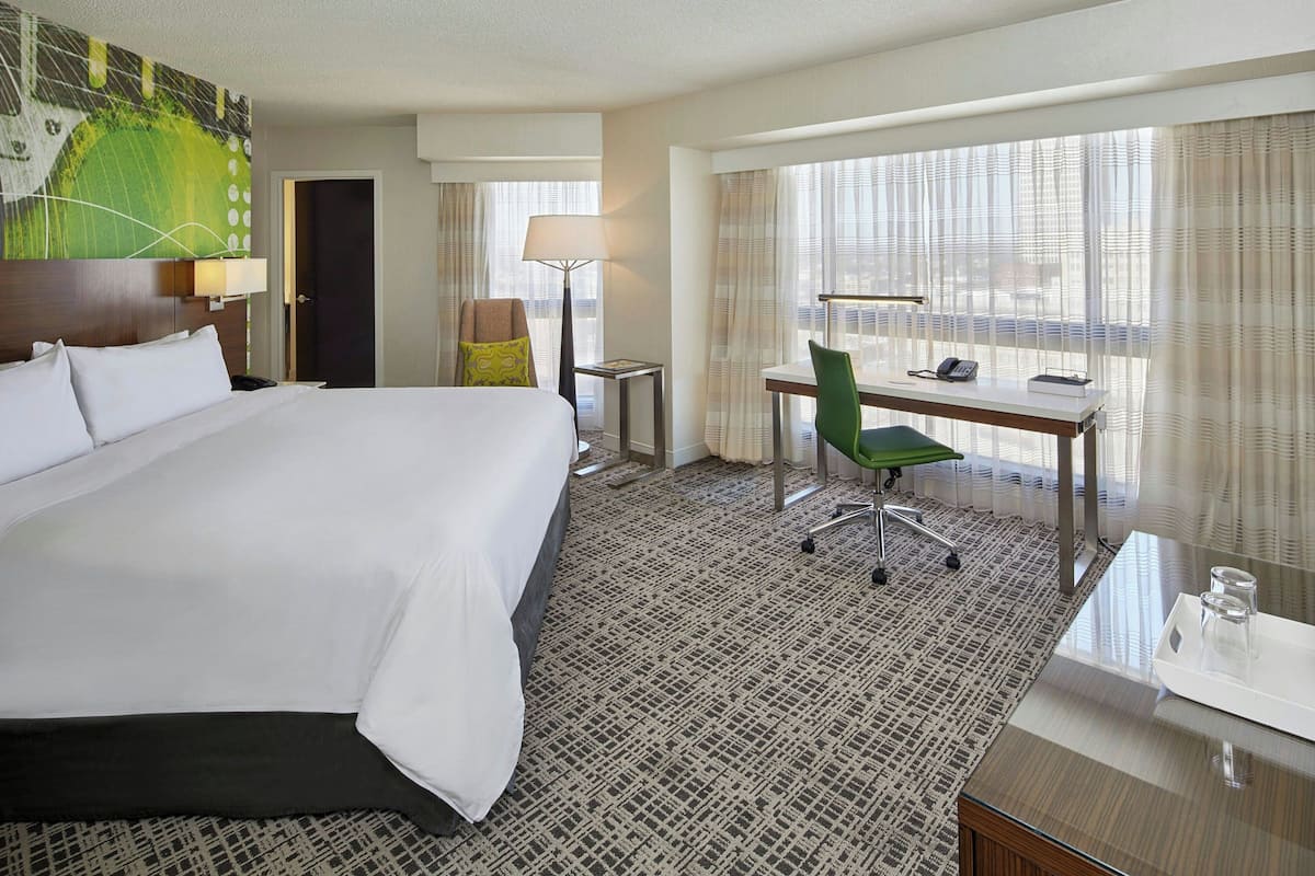 Habitación de hotel con una cama grande, un escritorio con silla, una lámpara y vistas a la ciudad desde la ventana.