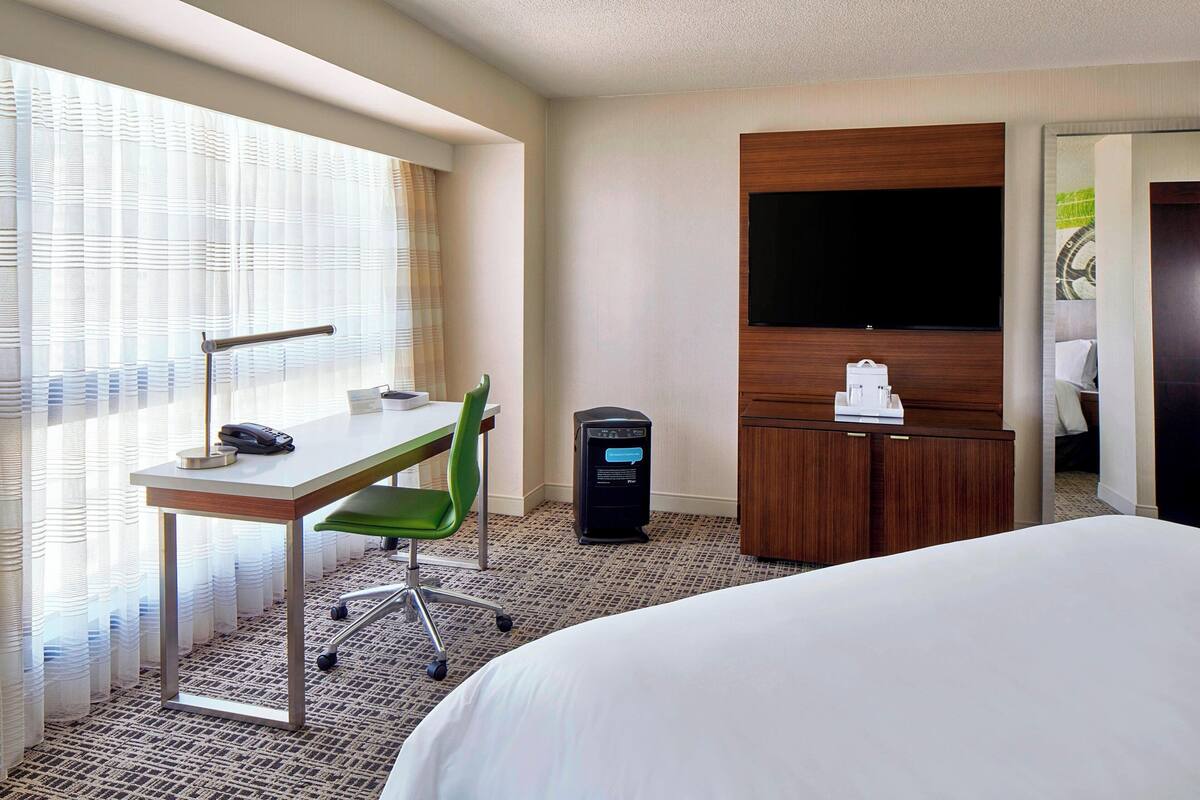 Una habitación de hotel con cama, escritorio con silla, computadora, televisor en la pared y armario.