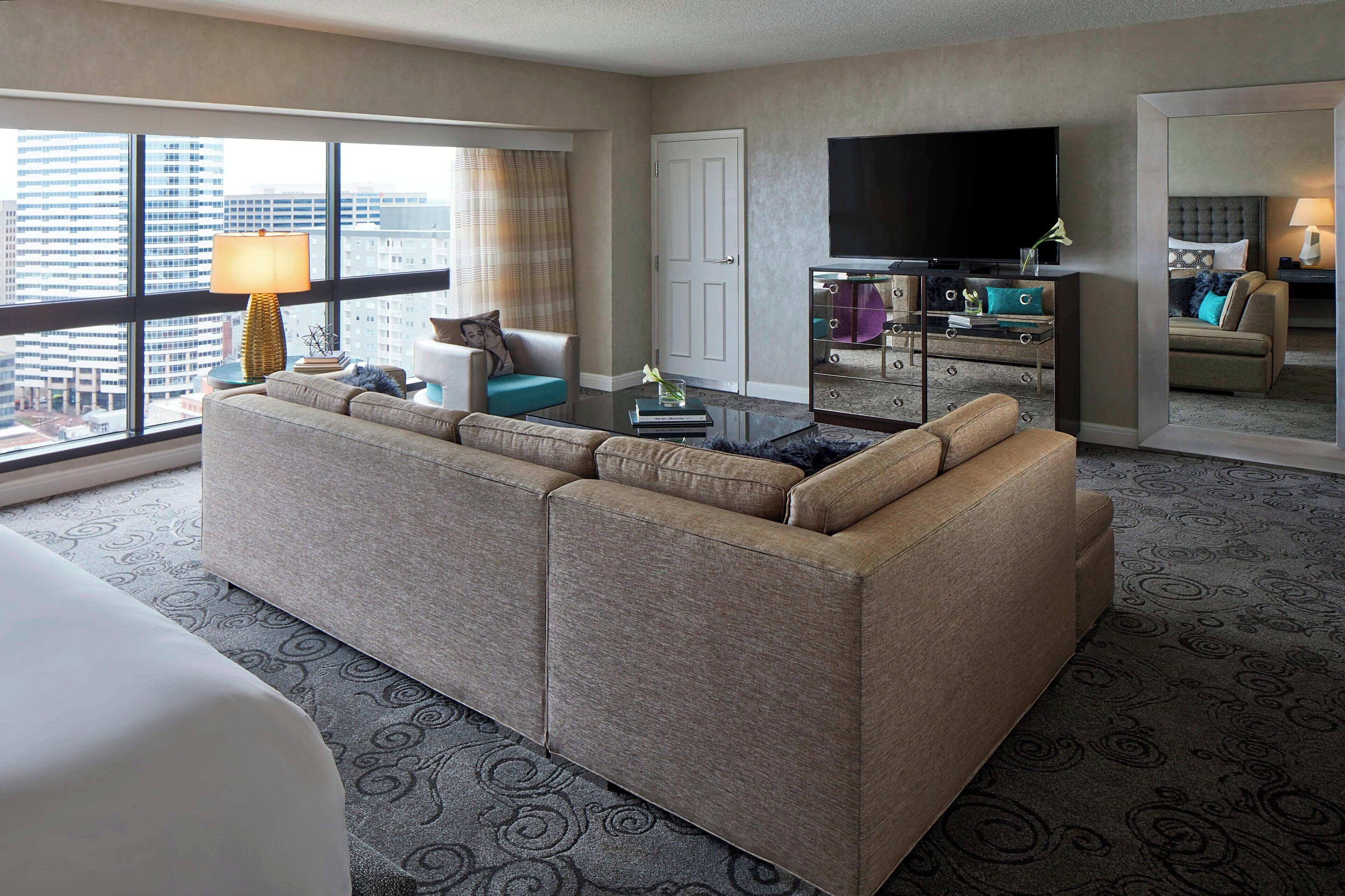 A hotel room with a large sectional sofa, a flat-screen TV, a coffee table, and a city view through large windows.