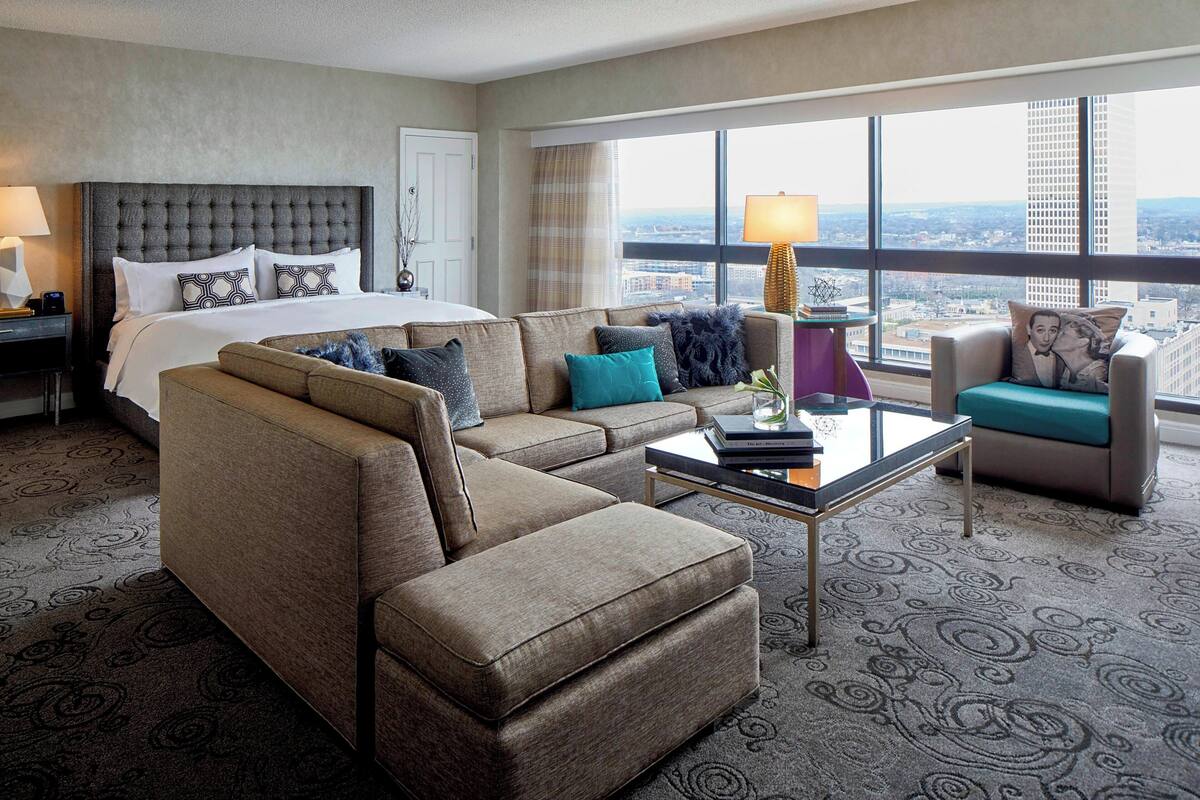 Habitación de hotel con una cama grande, un sofá, una mesa de centro y vistas de la ciudad a través de amplios ventanales.