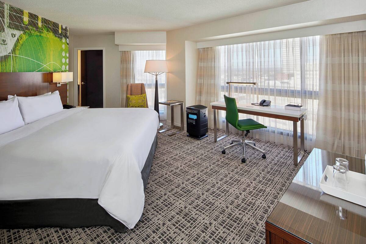 Habitación de hotel con una cama grande, un escritorio con computadora, una silla y una ventana con cortinas.