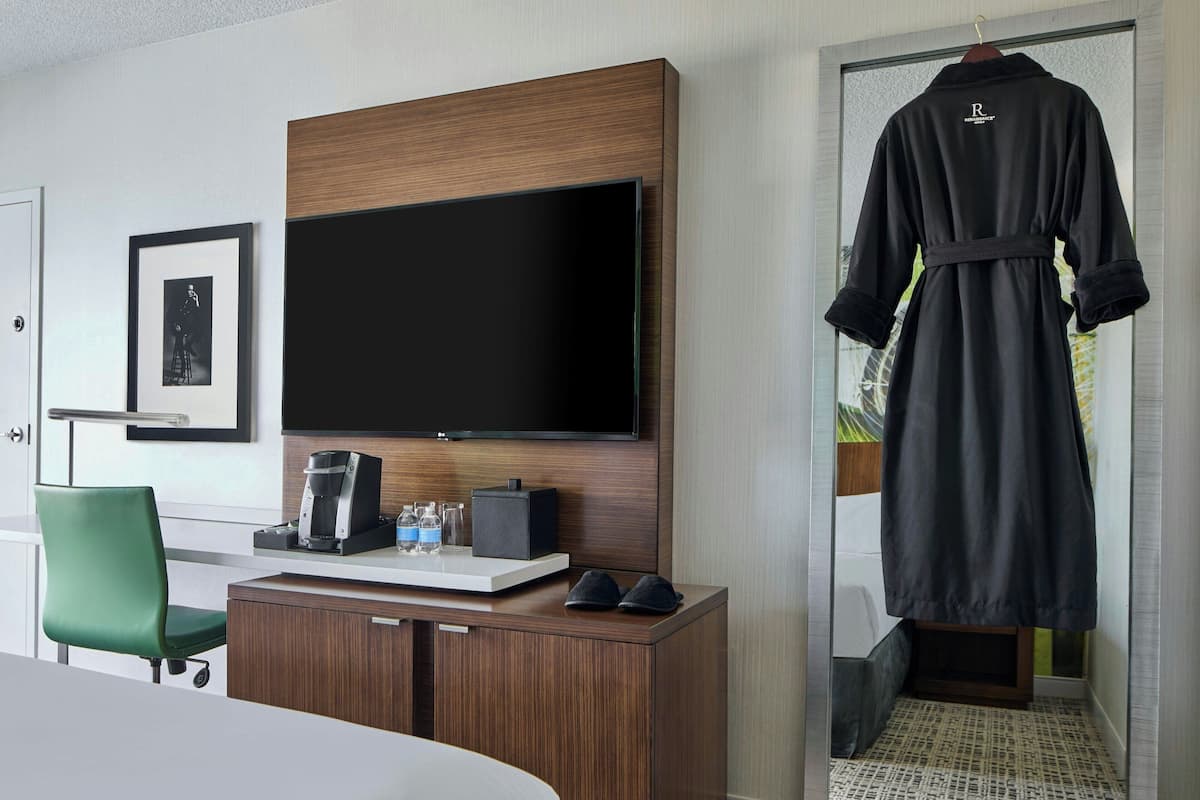 Habitación de hotel con televisor de pantalla plana, minibar, una silla verde y una bata colgada en la puerta.