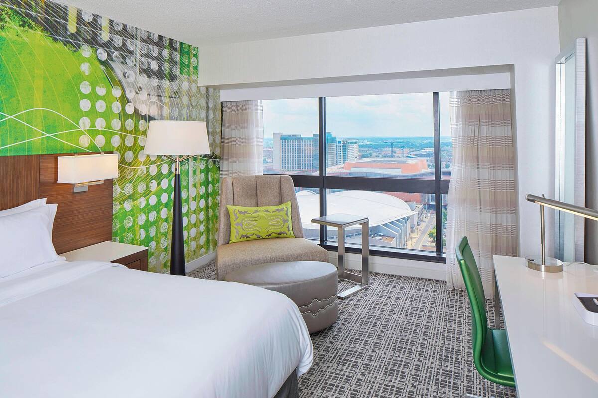 Habitación de hotel con cama, una silla, una mesita y vistas a un paisaje urbano a través de amplios ventanales.