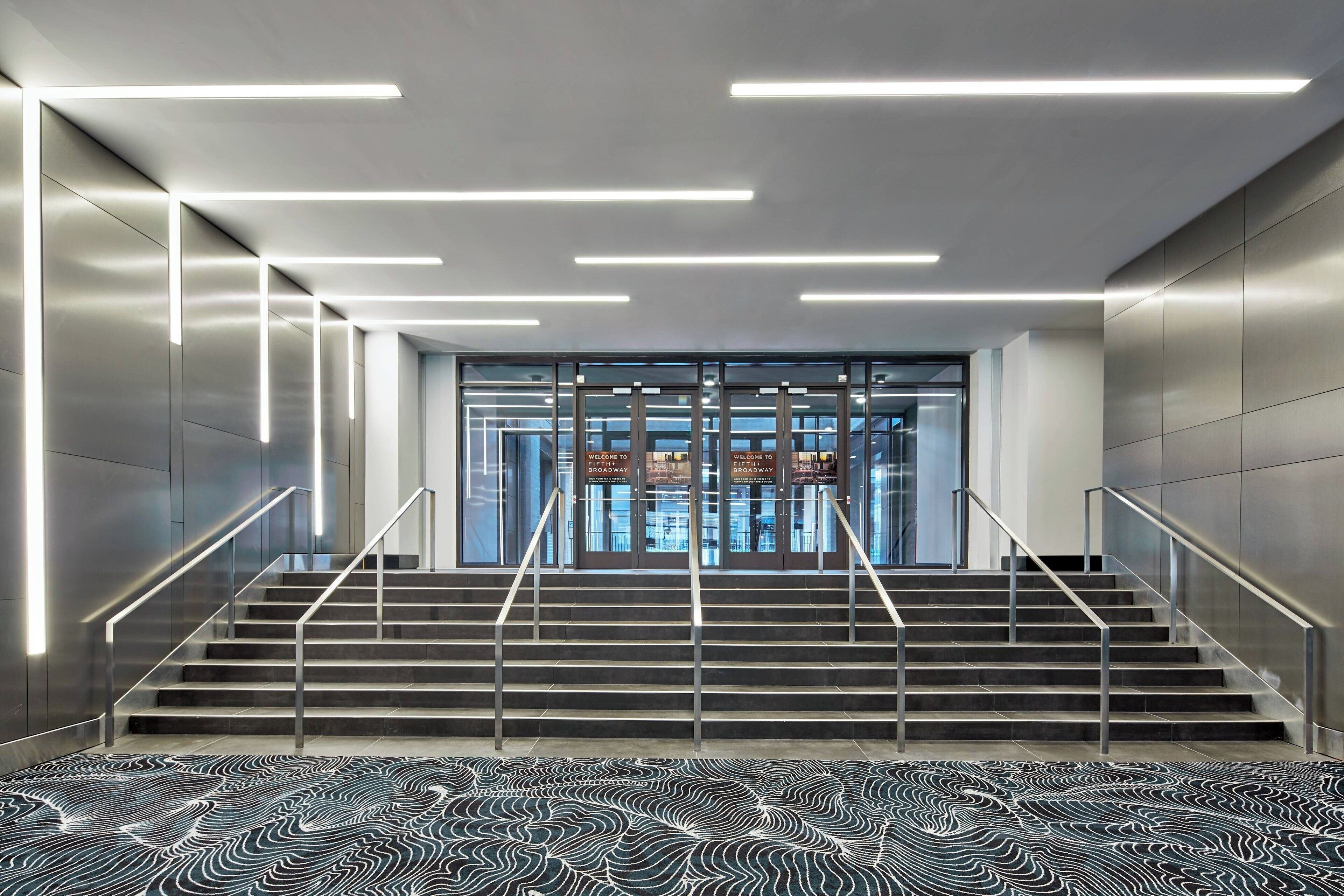 A modern staircase with metal railings and a patterned floor, leading to a glass door entrance with signage.