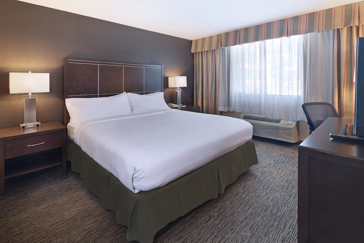 A hotel room with a large bed, two bedside tables with lamps, a desk, and a window with curtains.