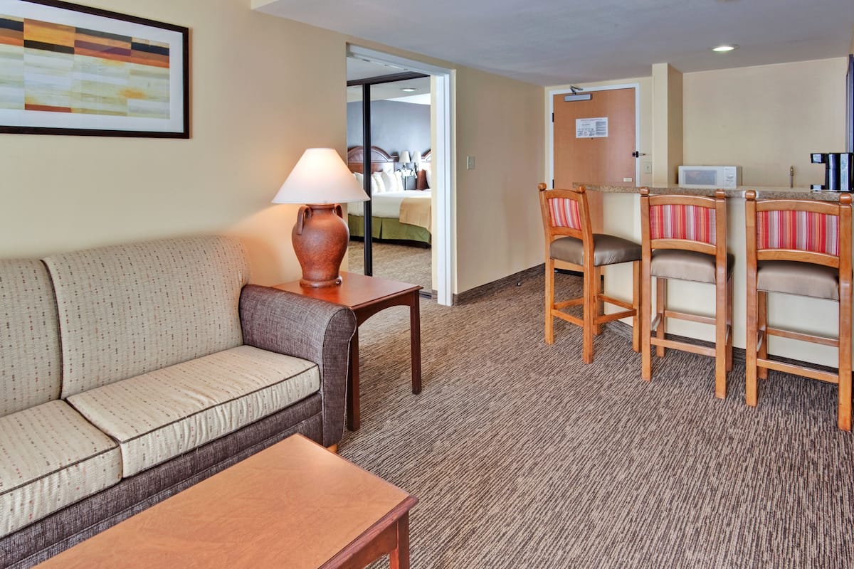A hotel room with a sofa, a small table with a lamp, a kitchenette with a microwave, and a bed visible through an open door.
